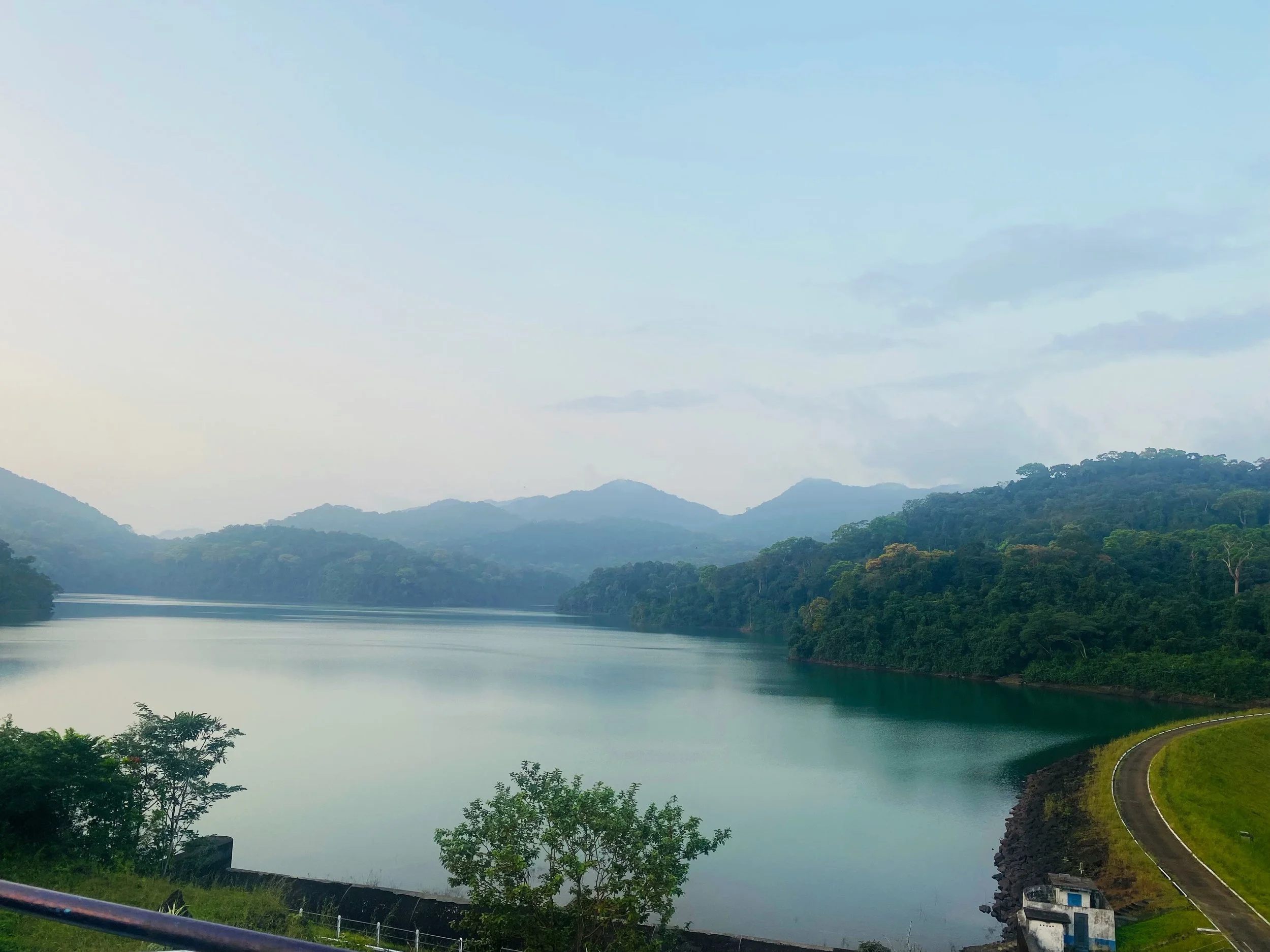 A scenic view of a calm river or lake surrounded by green hills and trees, with a cloudy sky overhead and a winding road along the shoreline.