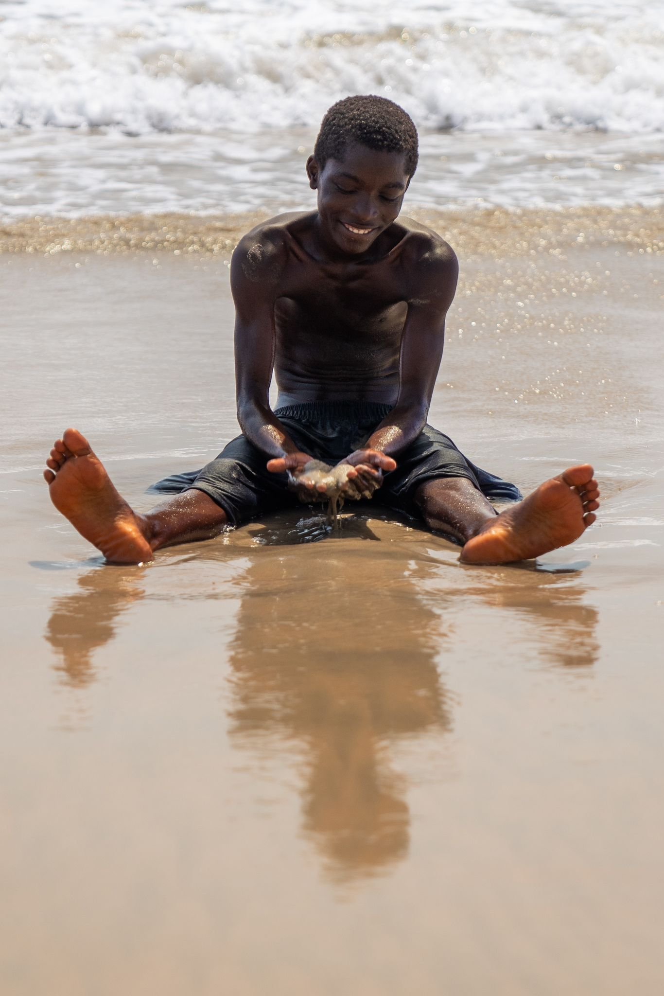 A young man sitting on the beach near the water with his legs spread out, holding a handful of seashells and smiling.