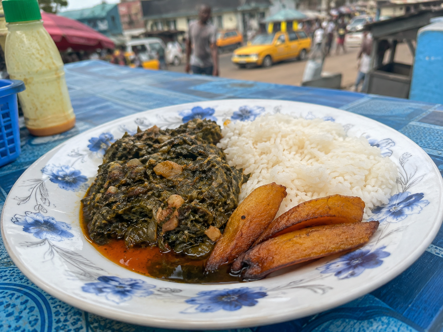 Plate of rice, cooked spinach stew, and fried plantains on a table outdoors with marketplace in background.