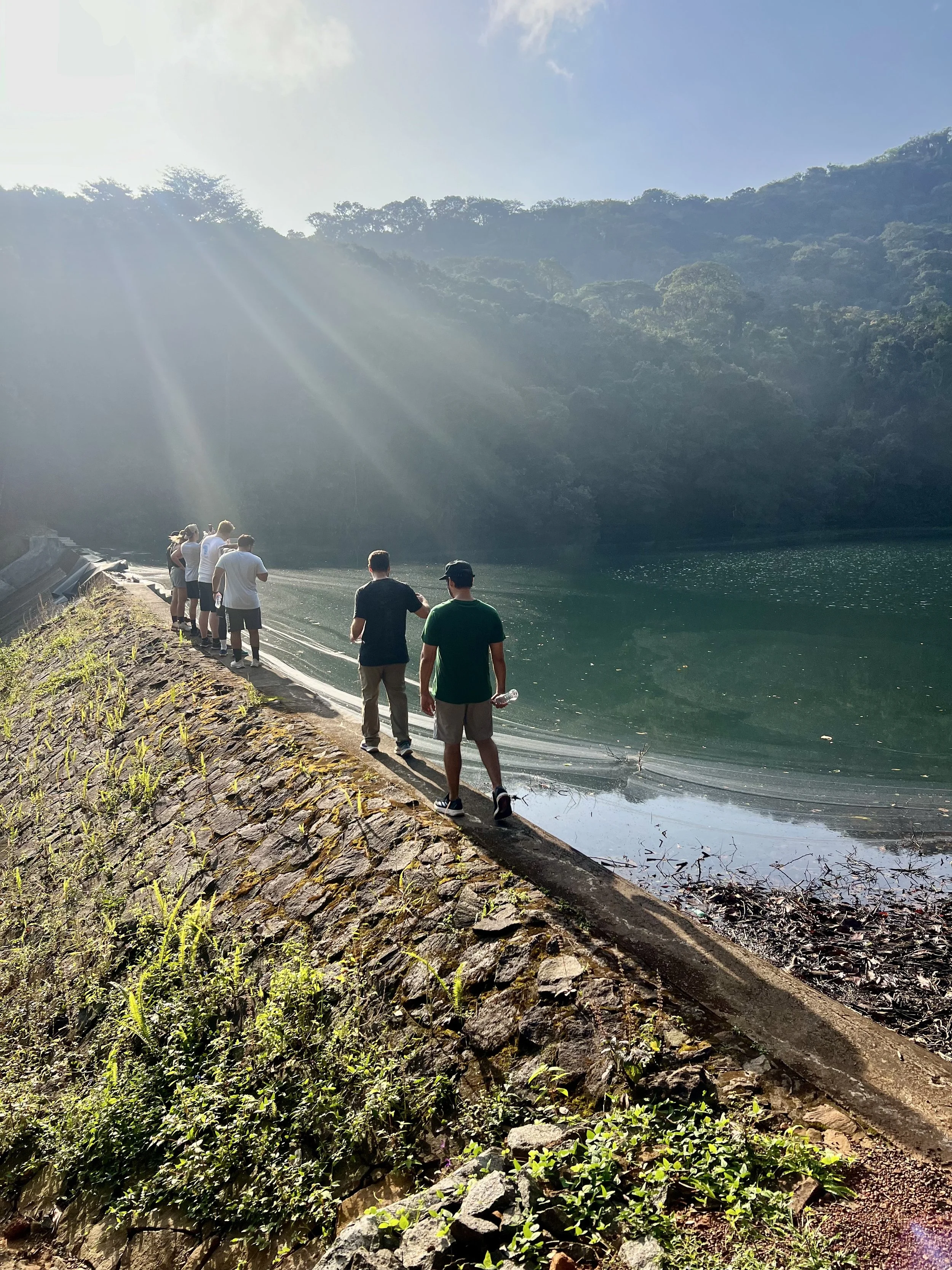 Group of people walking along a stone-lined path beside a body of water, with a forested mountain and blue sky in the background.