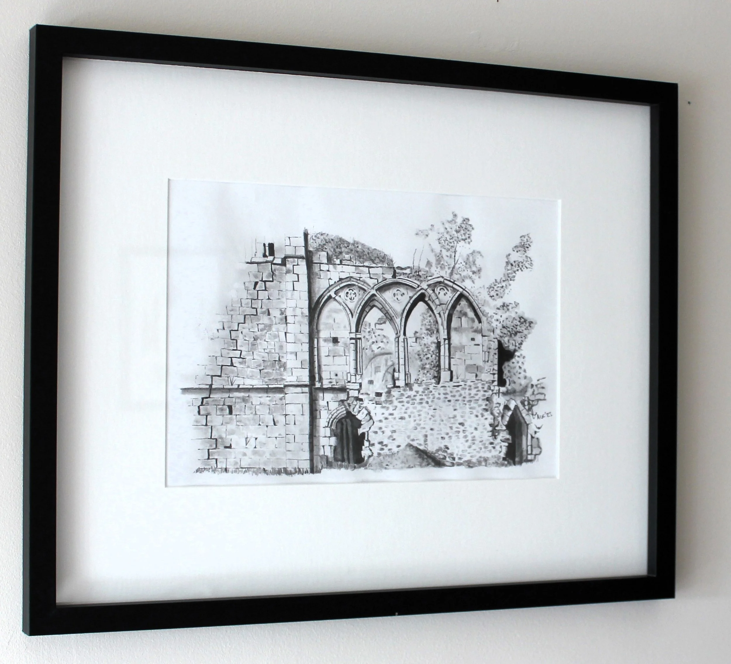 Black frame photograph of ancient stone ruins with arches and a partially collapsed wall, set outdoors with trees in the background.