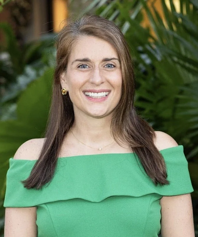 A woman with long brown hair, wearing a green off-the-shoulder dress, gold earrings, and a small necklace, smiling outdoors with green foliage in the background.