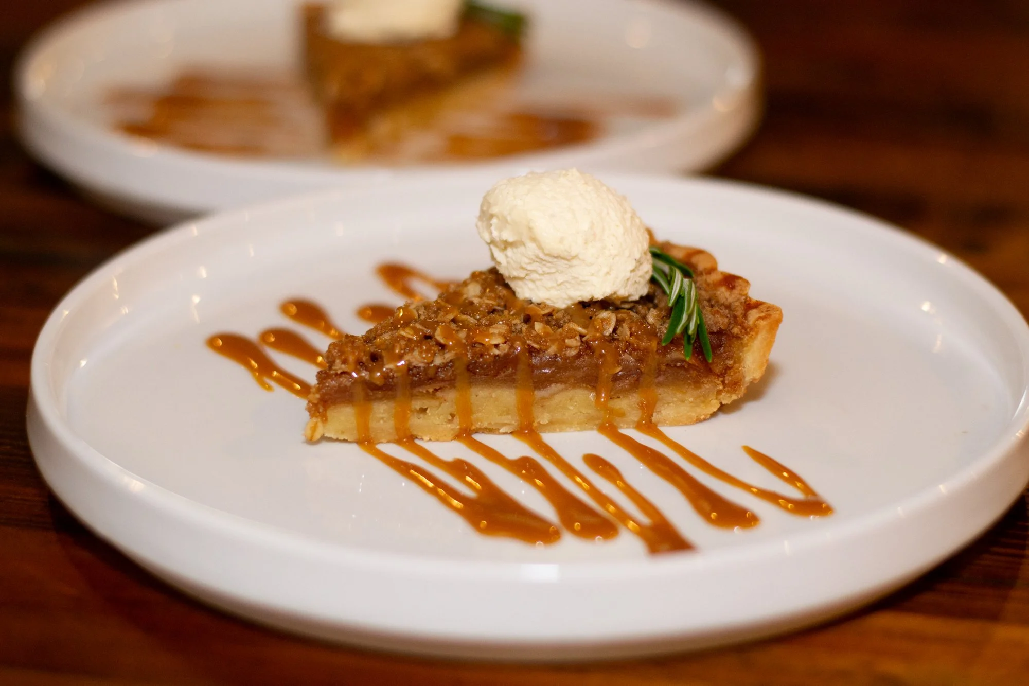 A slice of pecan pie topped with whipped cream on a white plate, drizzled with caramel sauce and garnished with a sprig of rosemary, with more pie on a separate plate in the background.
