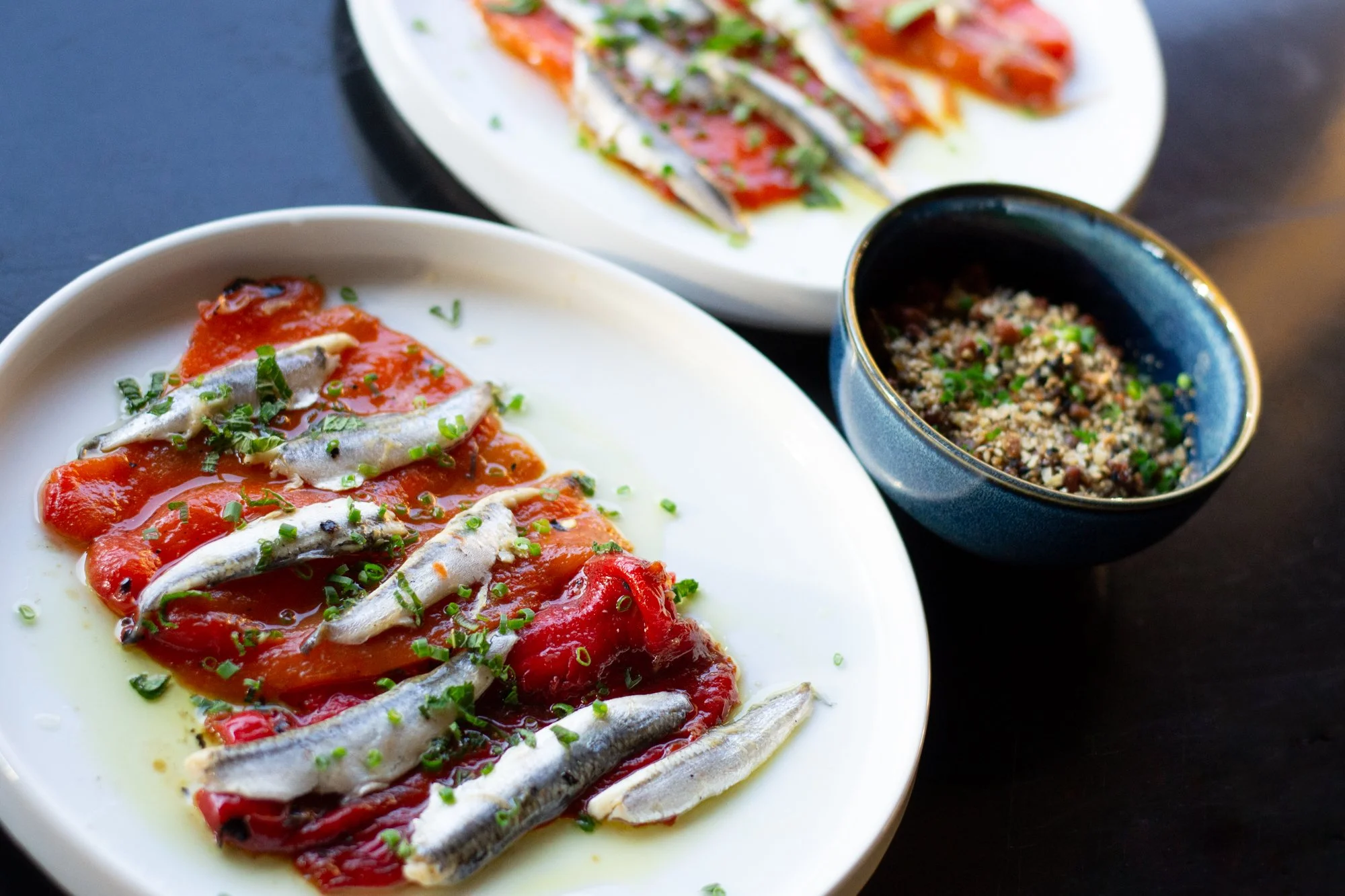 Plate of red sauce with small fish garnished with chopped green herbs, a bowl of mixed grains garnished with herbs, and a dish of sliced tomatoes with fish and herbs on top.