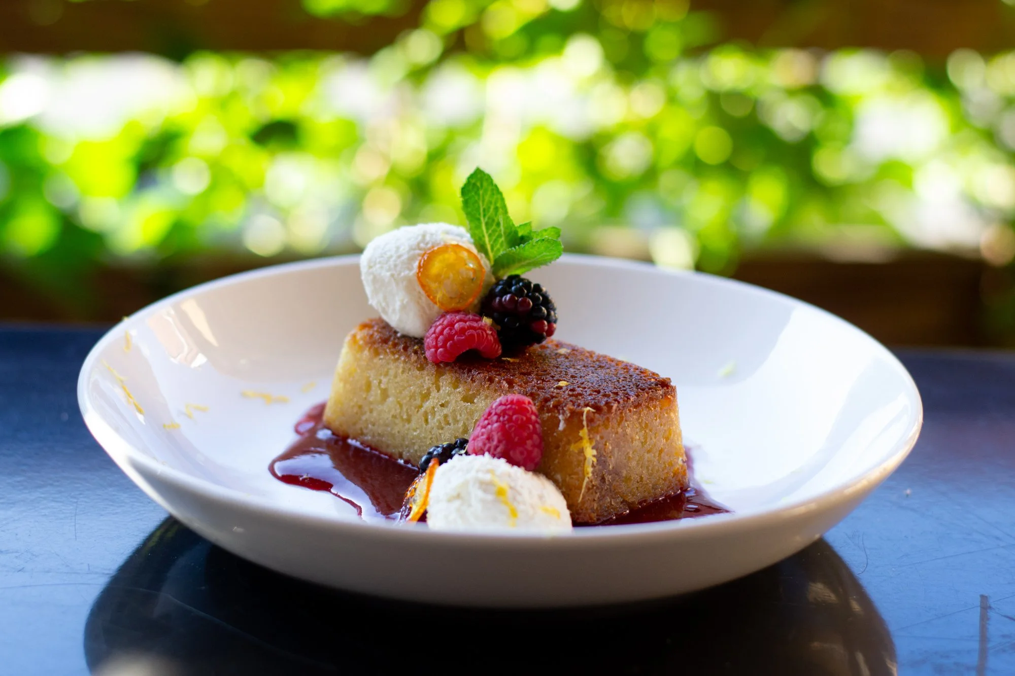A slice of cheesecake garnished with fresh berries, a dollop of whipped cream, and mint leaves on a white plate, with a blurred green leafy background.