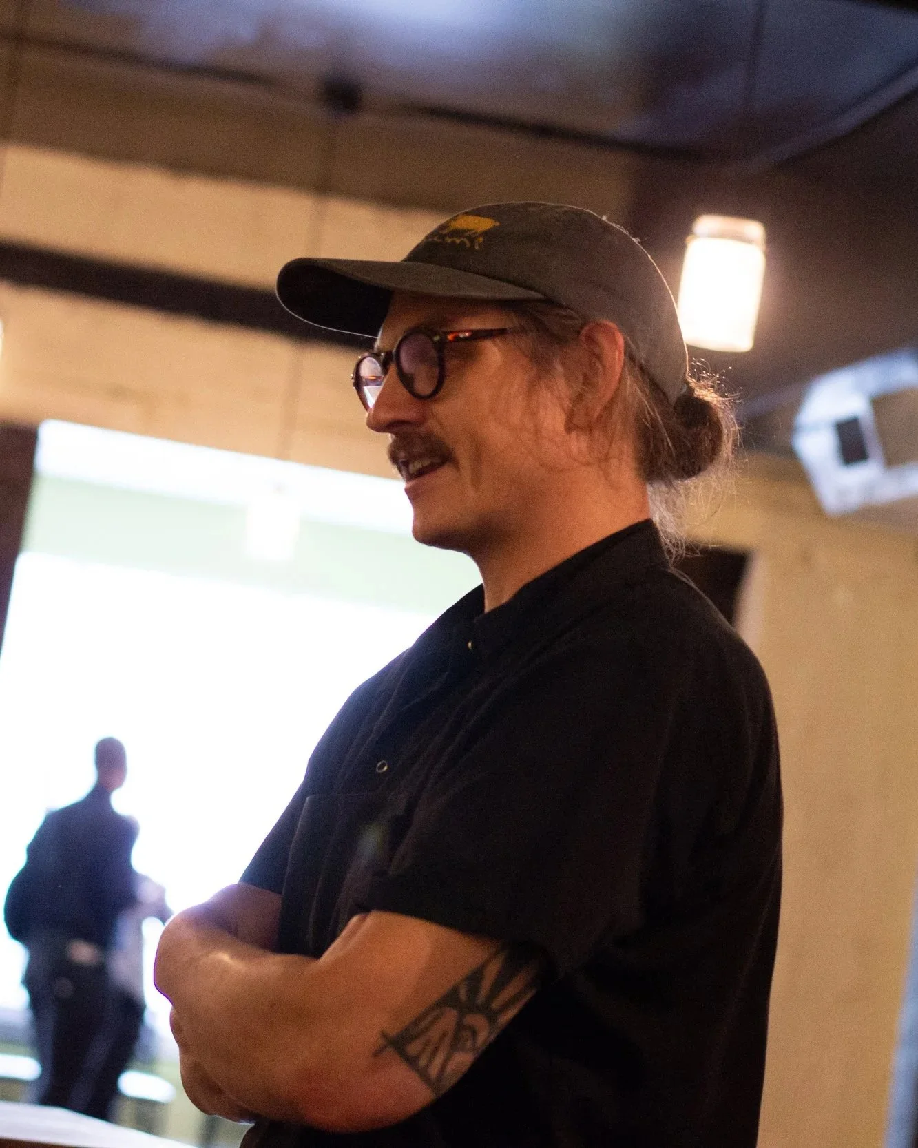 A man with glasses and a black cap, with long hair tied in a bun, is standing with his arms crossed in a dimly lit room, with a blurred background of other people and ambient lighting.
