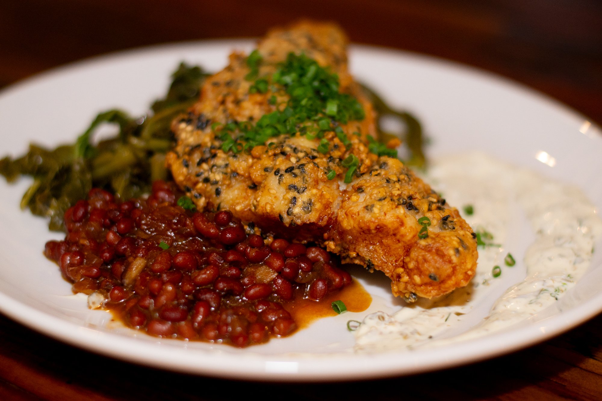Plate with baked fish topped with green herbs, served with red beans, green vegetables, and a creamy white sauce.