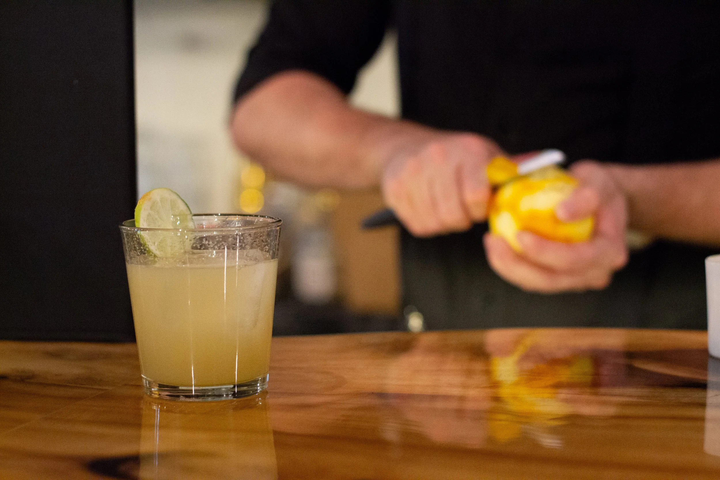 A hand holding a lemon, preparing a lemon beverage in the background with a glass of lemonade with lime wedge on a wooden surface.