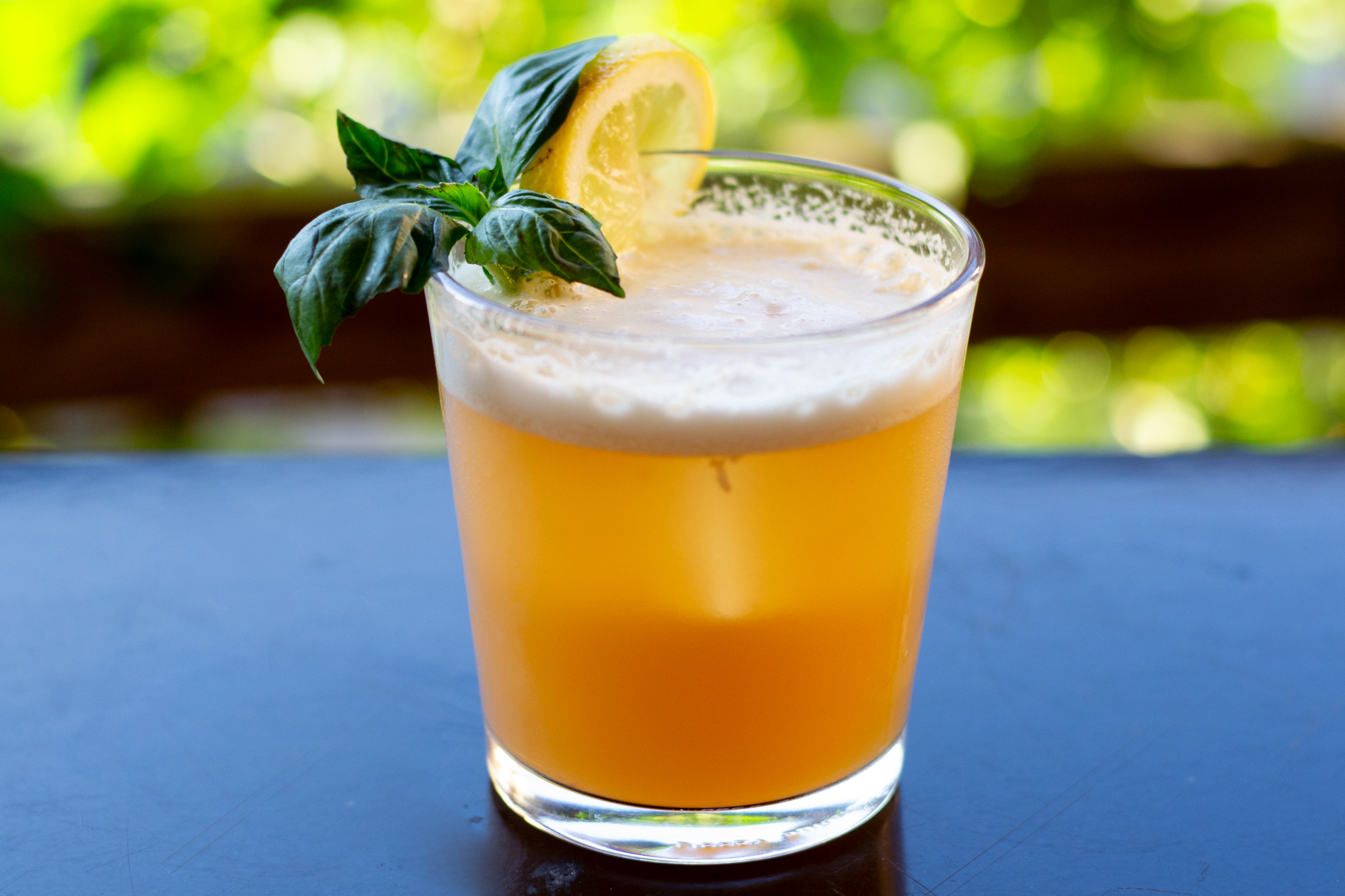 A glass of orange-colored beverage garnished with a slice of lemon and fresh green basil leaves, with a blurred green outdoor background.