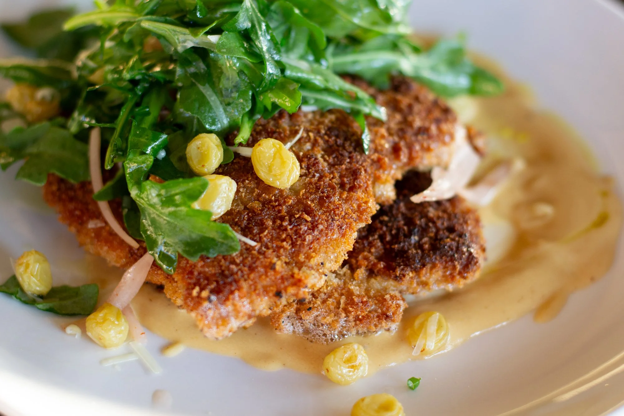 Close-up of a fried pork cutlet topped with arugula salad,  pickled raisins, and a creamy sauce on a white plate.