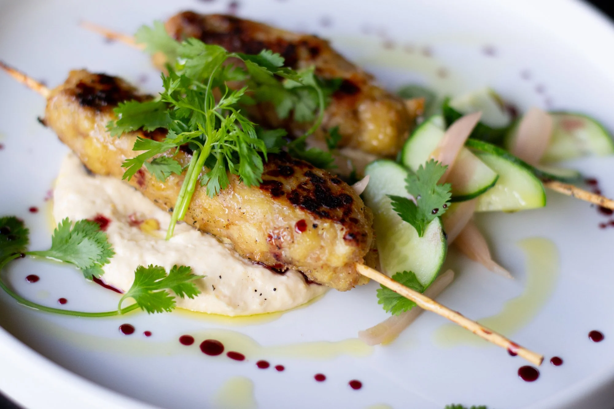 Close-up of grilled chicken skewer garnished with cilantro, served on a bed of hummus and accompanied by cucumber slices and sauce with red drizzles.