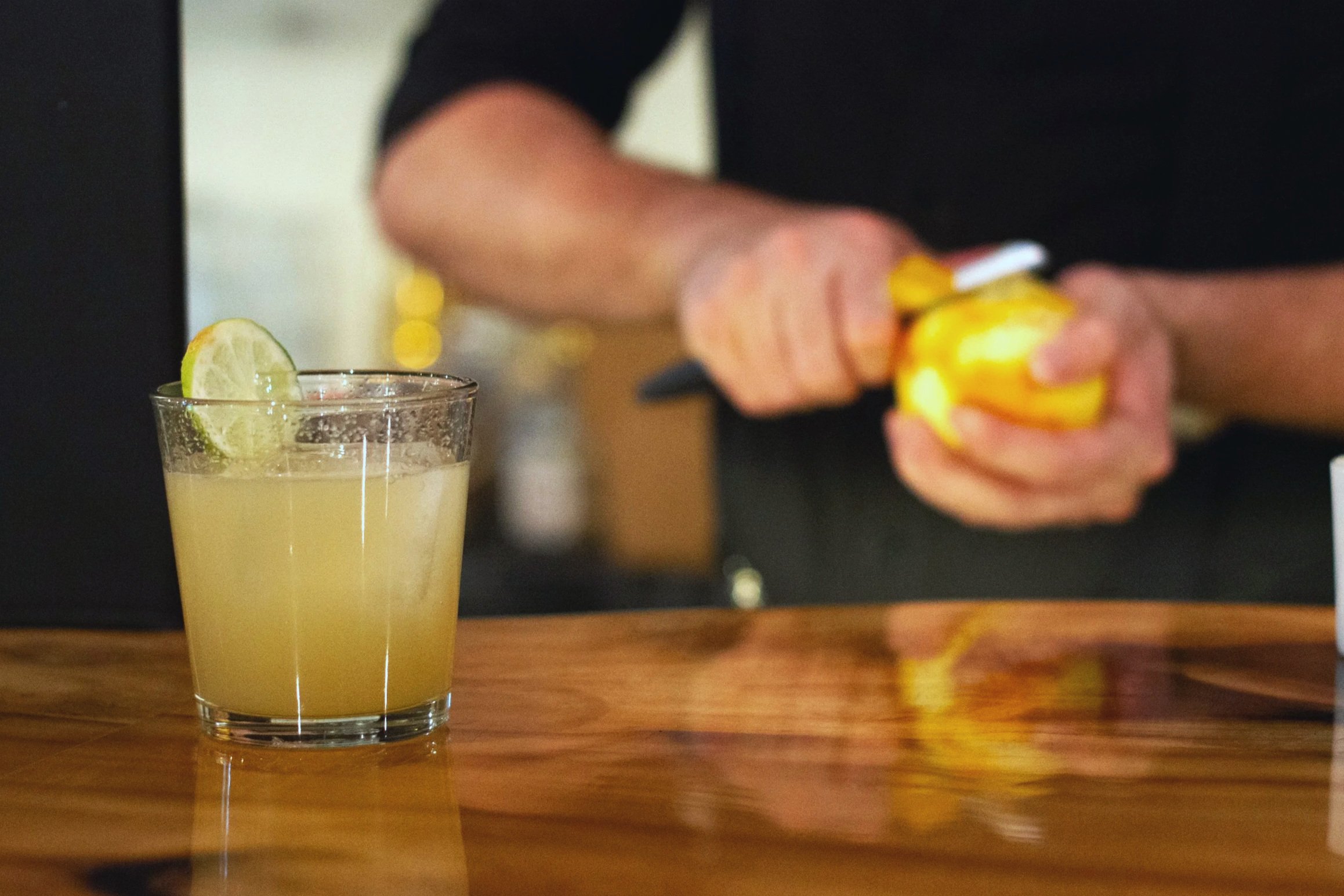 A close-up of a cocktail glass filled with a light yellow beverage, garnished with a lime wedge on the rim, placed on a wooden bar or table.