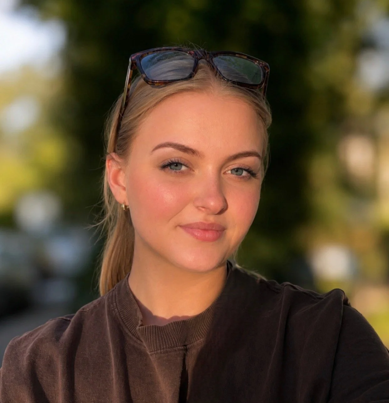 Close-up of a young woman with blonde hair, blue eyes, and sunglasses resting on her head, smiling slightly outdoors with a blurred background of trees and possibly cars.