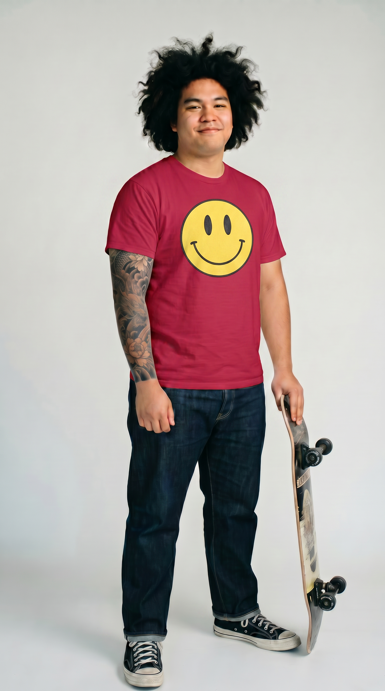 A young man with curly black hair and tattoos on his left arm, standing with a skateboard, wearing a red T-shirt with a yellow smiley face, dark jeans, and black Converse sneakers, smiling at the camera in front of a plain white background.