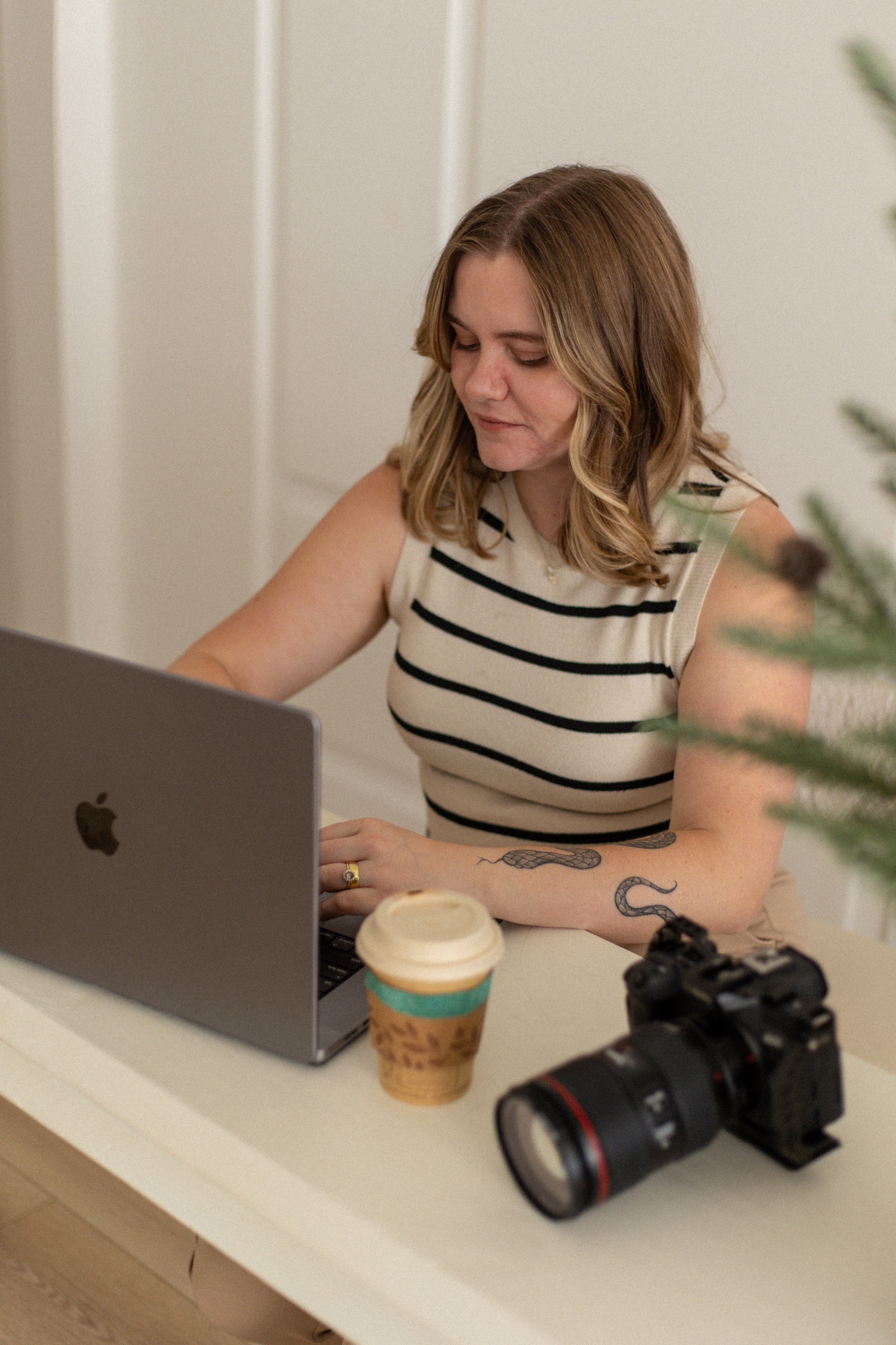 A woman with shoulder-length, wavy blonde hair, wearing a sleeveless, striped top, sitting at a desk with a laptop, a coffee cup, and camera equipment, in an indoor setting.