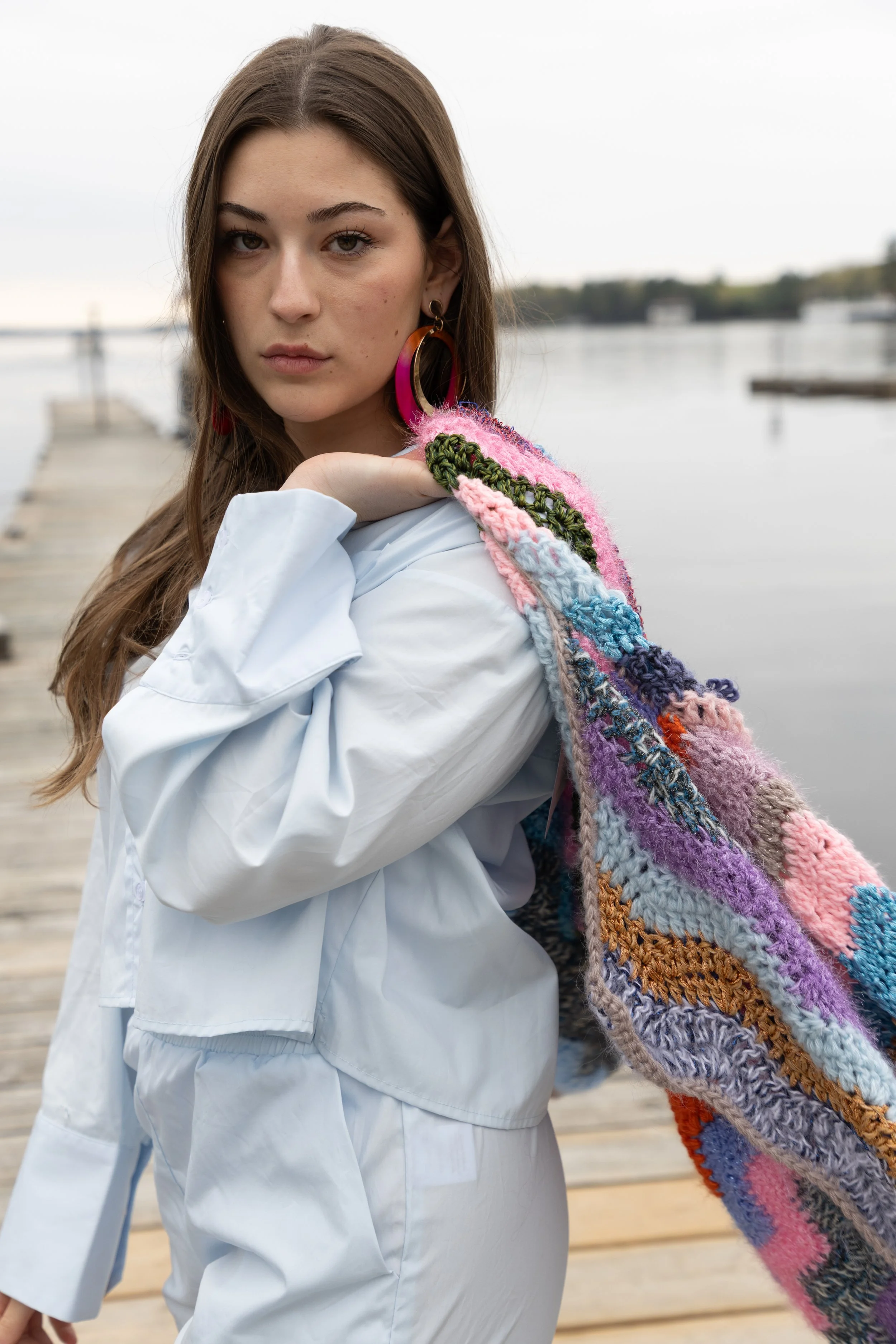 A young woman with brown hair and hoop earrings, wearing a white jacket and a colorful, knitted scarf, standing on a dock near water, looking at the camera with a serious expression.