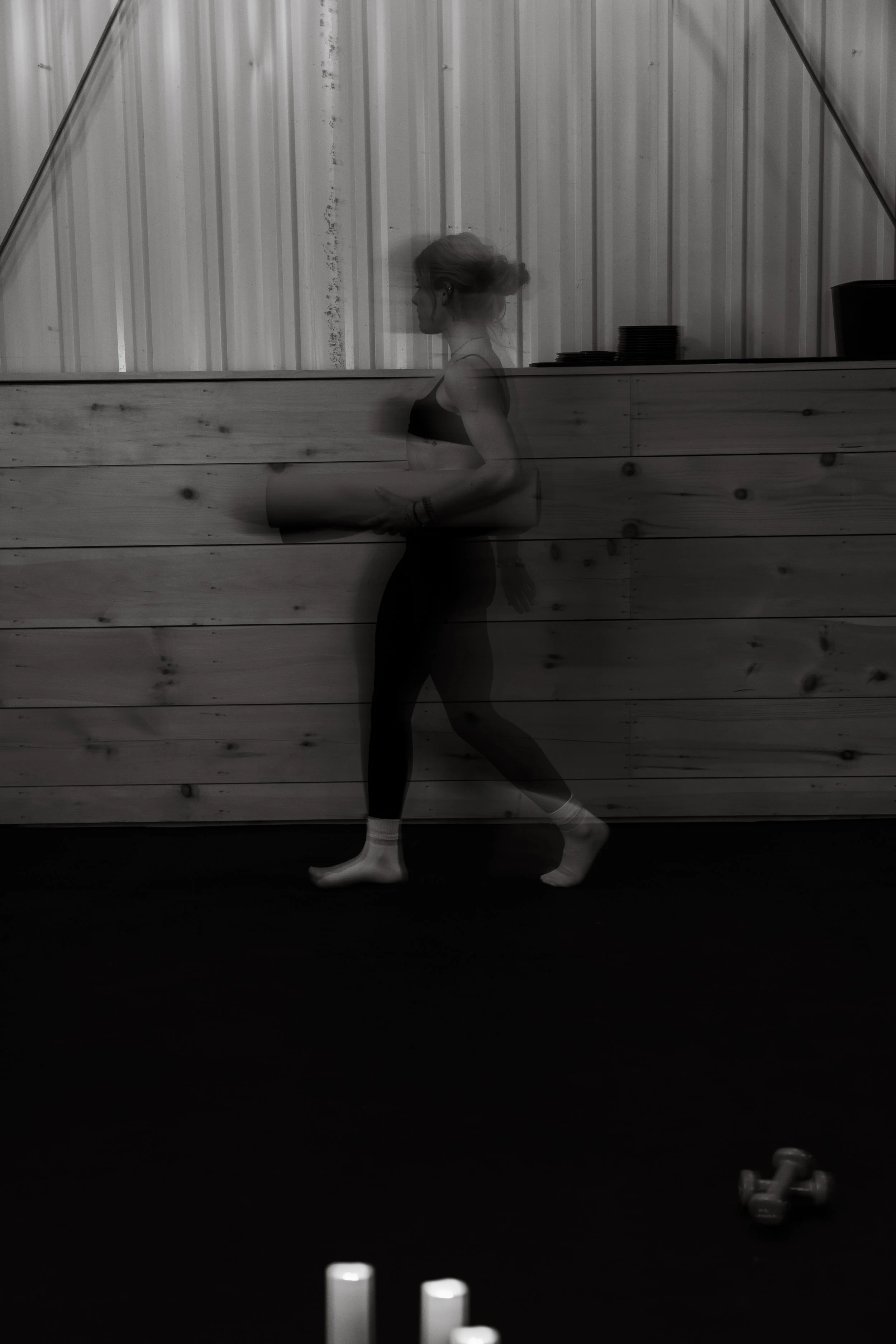 A woman in workout attire doing yoga in a gym with dark flooring, a wood-paneled wall, and stacked plates in the background. Exercise equipment is visible in the foreground.