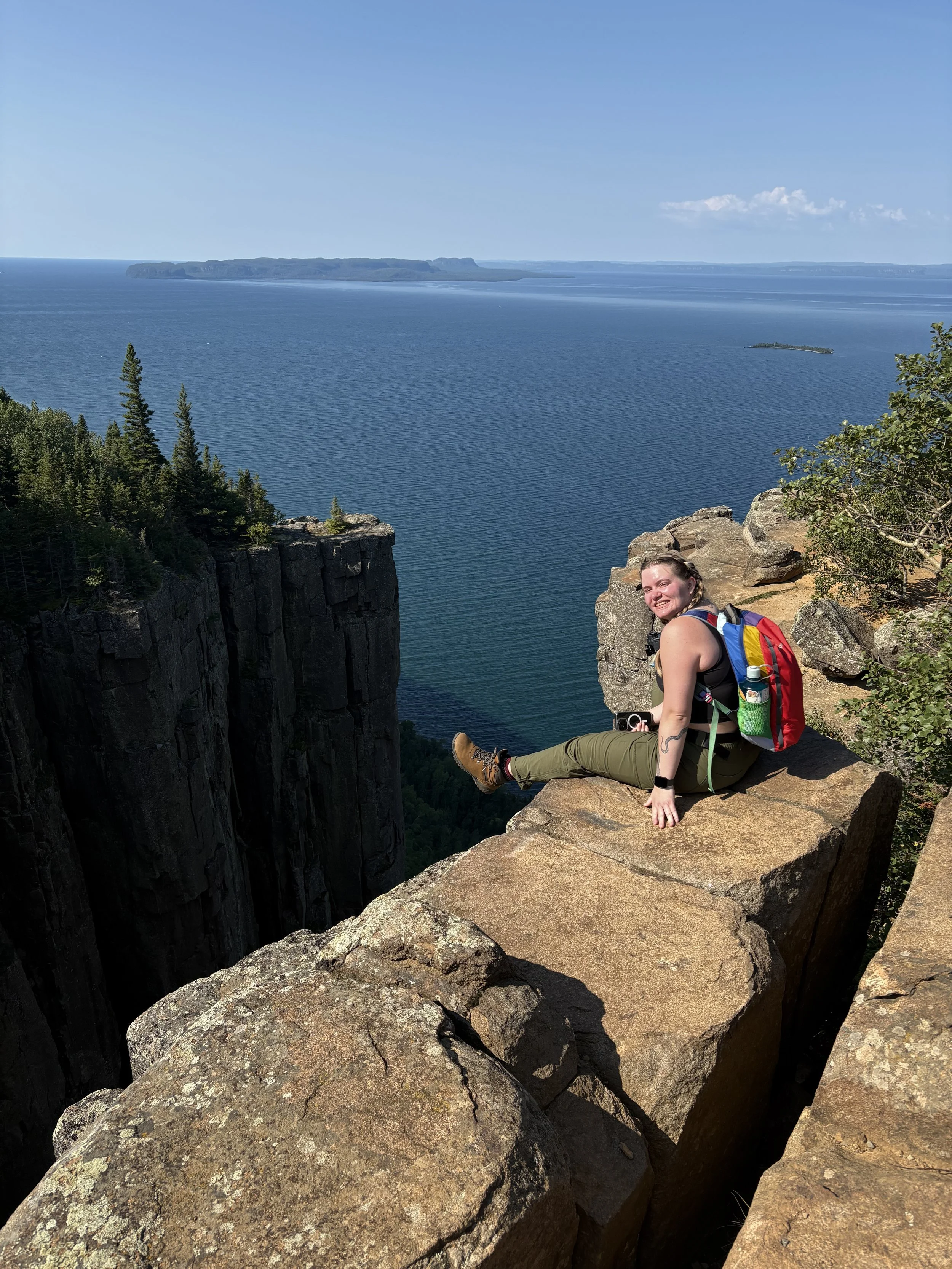 A smiling woman with a backpack sitting on a rocky overlook with a large body of water and distant land in the background.
