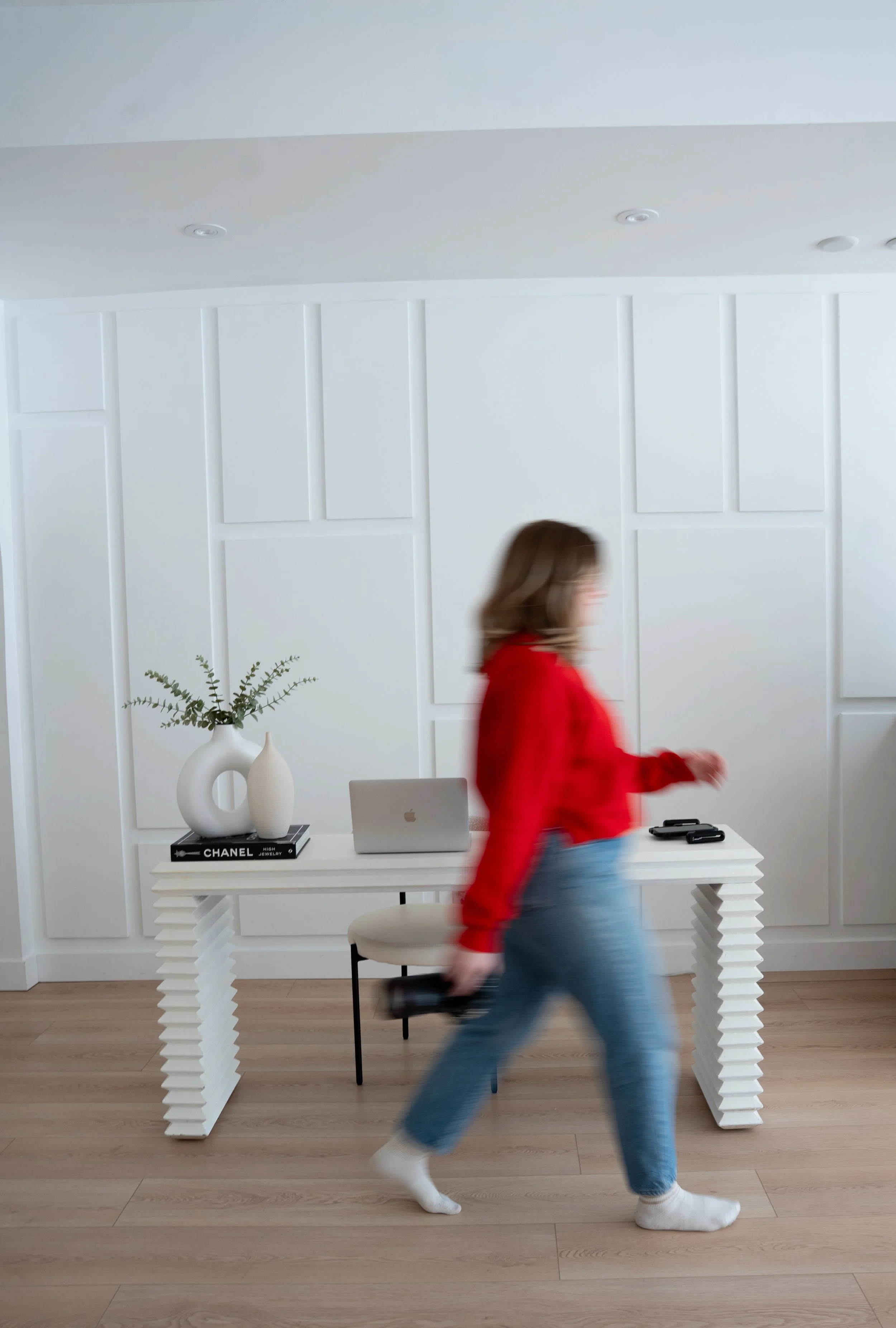 A woman in a red sweater and blue jeans walking briskly past a white table with decorative vases, a laptop, and cell phones, in a minimalist white room with wooden flooring.