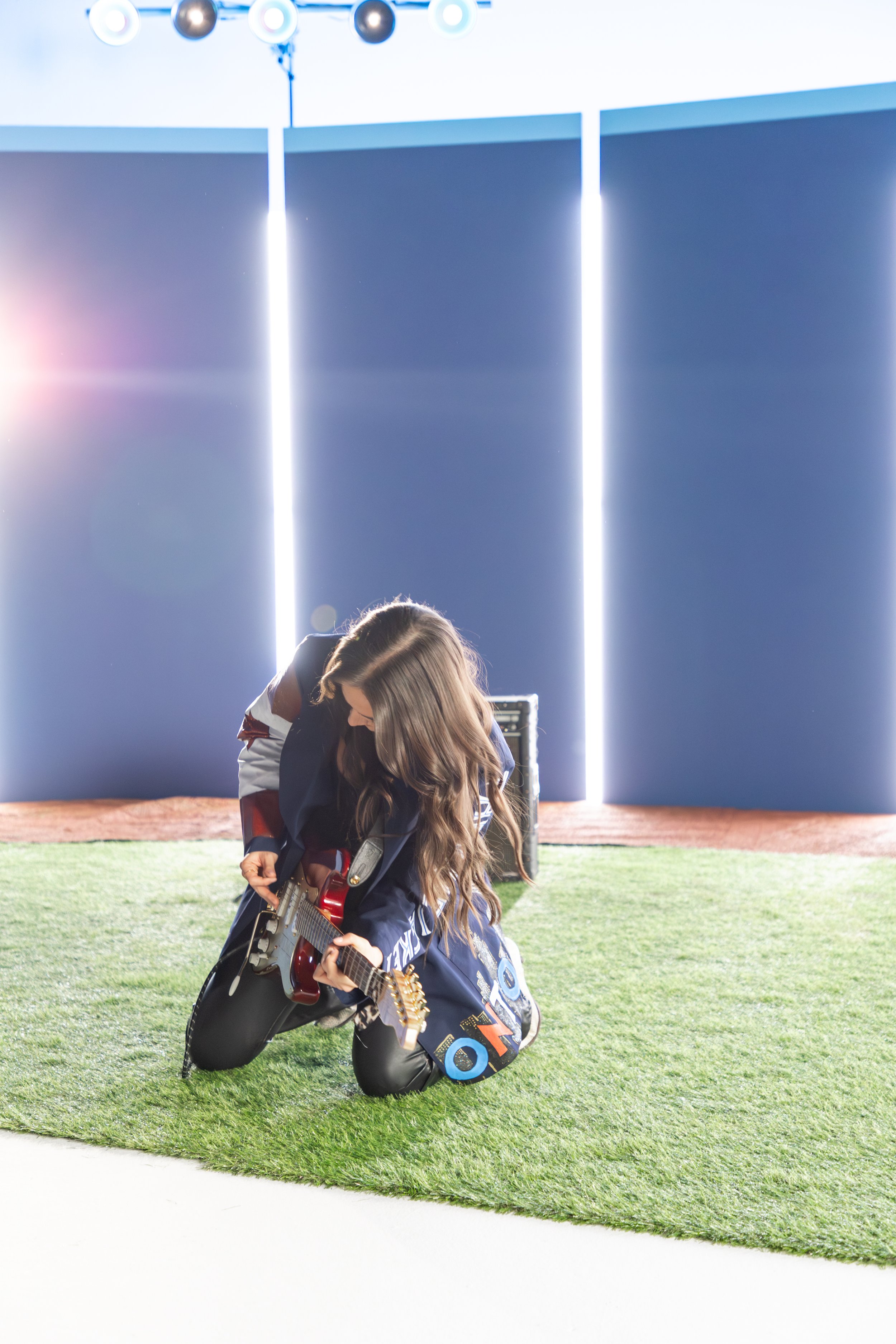 The artist JIMI kneeling on a baseball field playing an electric guitar during a performance, with a blue backdrop and stage lights overhead.