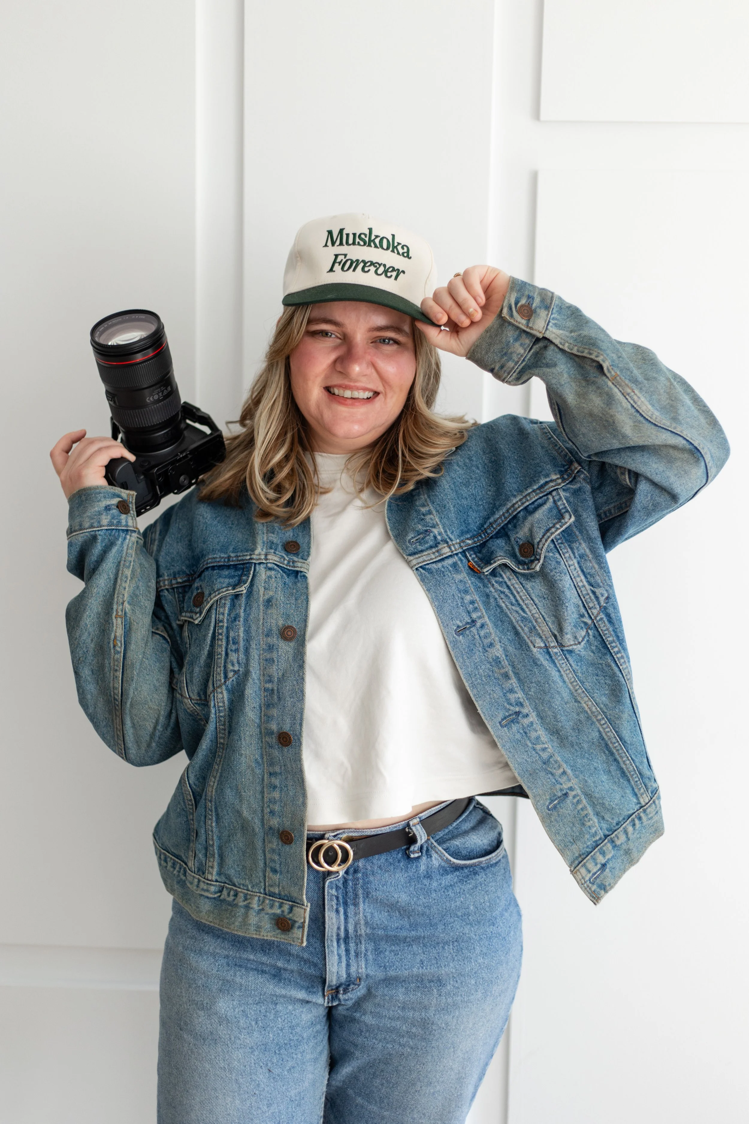 A woman wearing a denim jacket and jeans, holding a camera, smiling, and adjusting a white cap that says "Muskoka Forever" in green letters, standing against a white background.