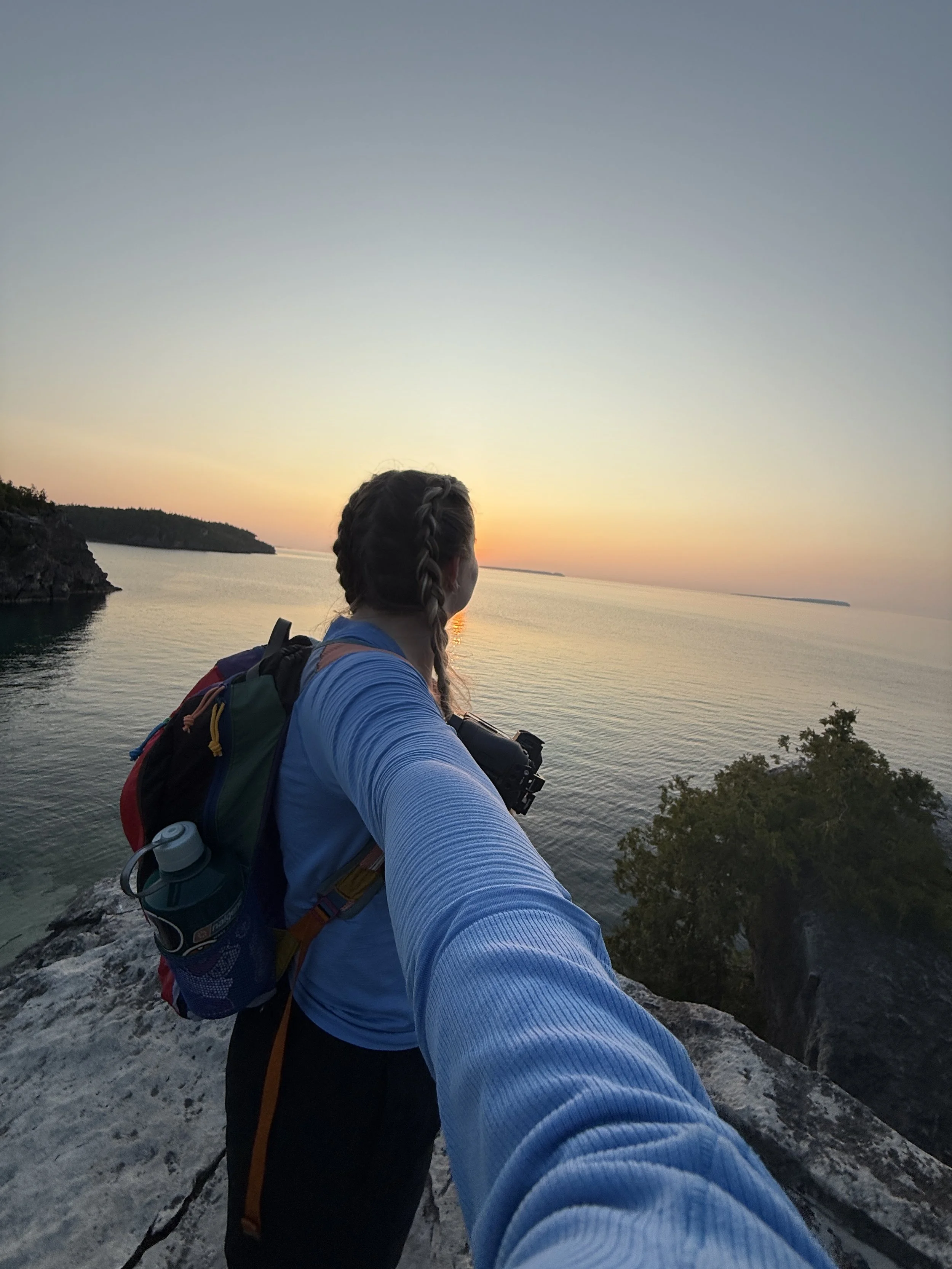 A person with braided hair and a backpack taking a selfie on a cliff overlooking a body of water at sunset.