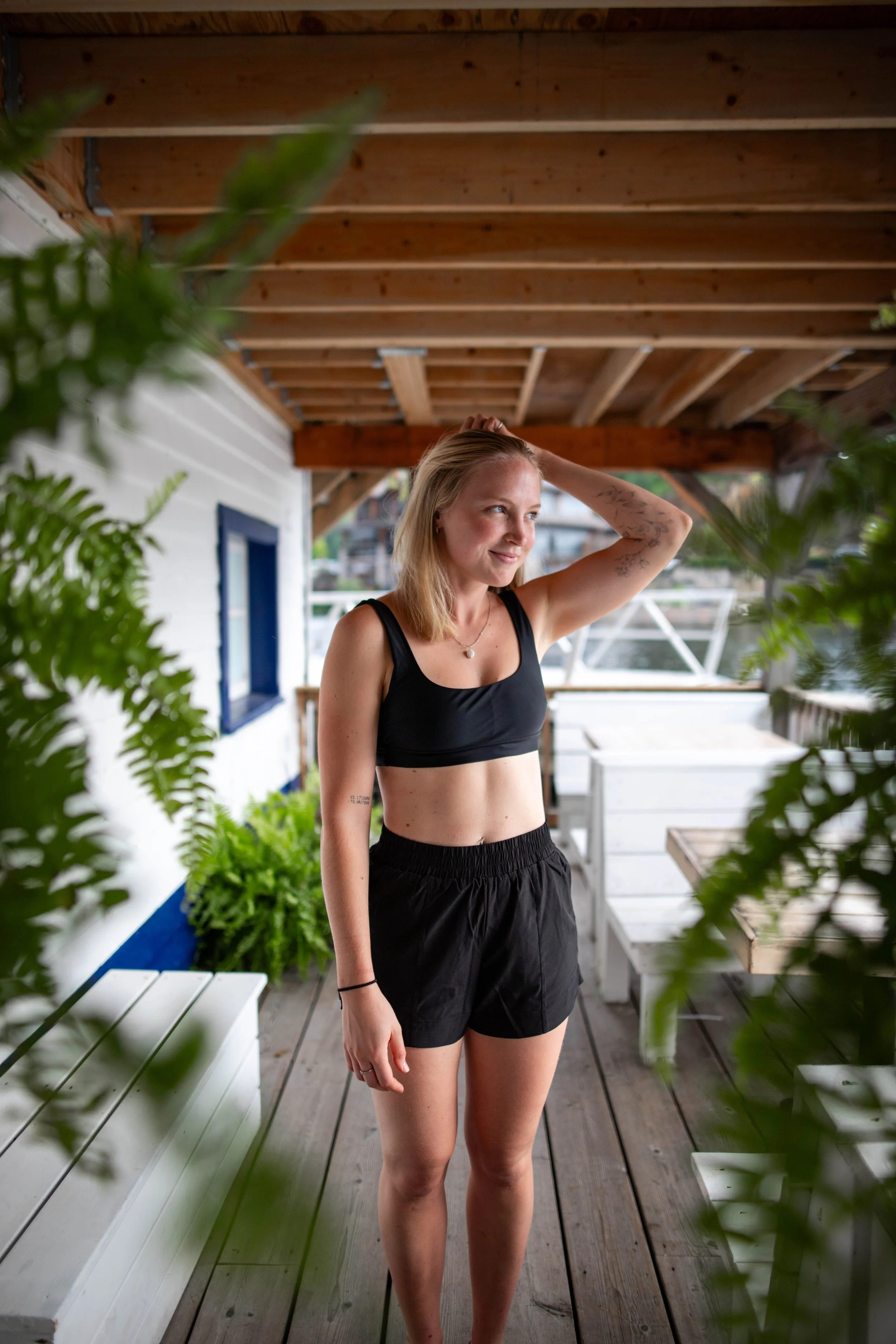 A woman standing on a wooden deck under a wooden roof, surrounded by green plants, wearing a black sports bra and black shorts, with blonde hair and a tattoo on her right arm, smiling and posing with her right hand on her head.