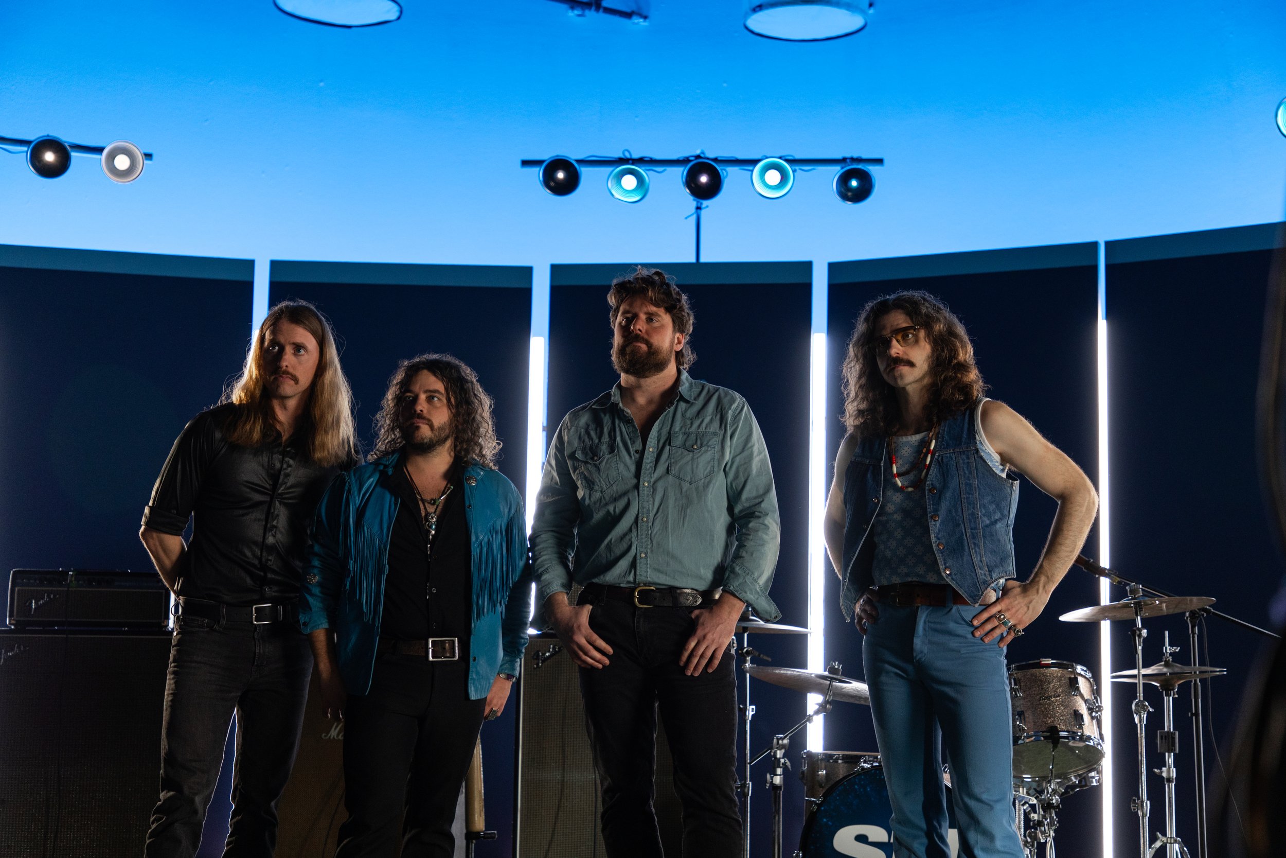 The Sheepdogs standing together in a music studio with stage lights above and musical instruments behind them.