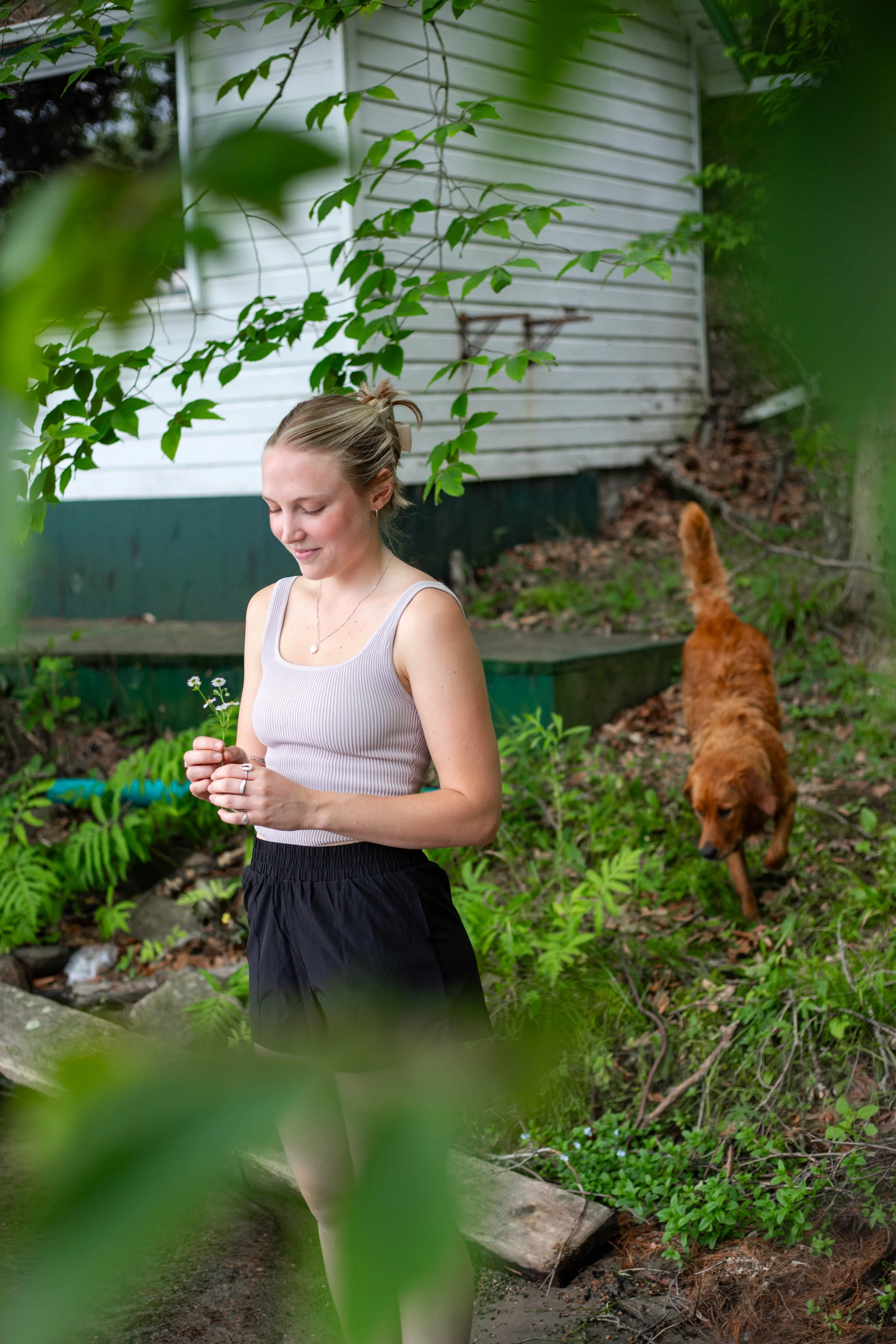 A young woman with blonde hair in a ponytail, wearing a light gray sleeveless top and black shorts, stands outdoors in a green, leafy garden next to a dog with reddish-brown fur. She is holding small flowers and smiling softly with her eyes closed, surrounded by plants and trees, with a white house in the background.
