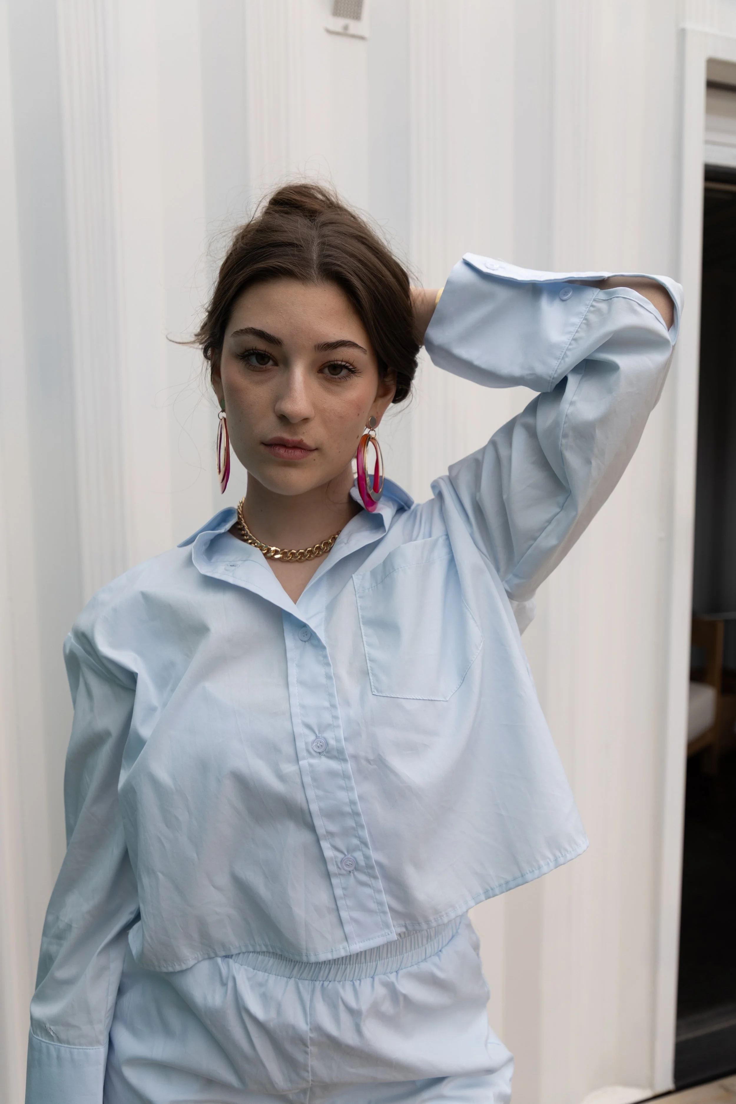 Young woman with brown hair wearing a white button-up shirt, large colorful earrings, and a gold chain necklace, standing indoors against a white and beige wall, touching her head with her right hand.