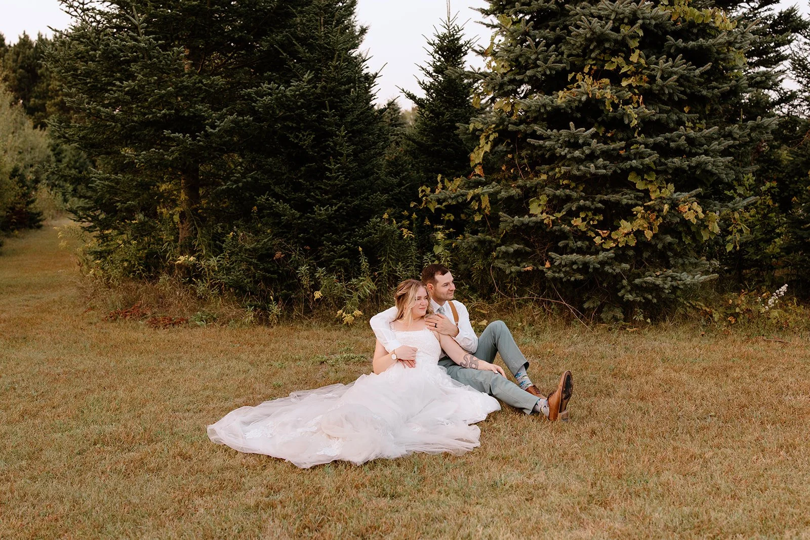 A couple dressed in wedding attire sitting on the grass in front of pine trees, with the bride in a white wedding gown and the groom in a white shirt and gray pants, outdoors during daytime.