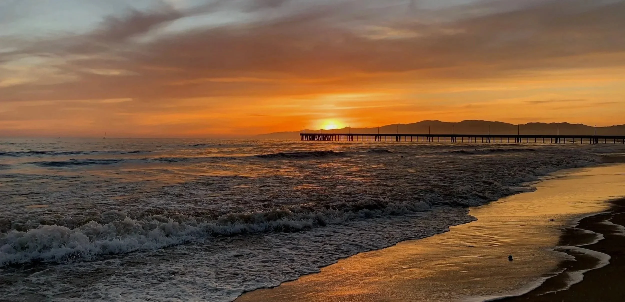 Sunset over the ocean at Venice Beach with a pier extending into the water and a sailboat in the distance, with the Santa Monica mountains on the horizon and waves crashing on the shoreline.