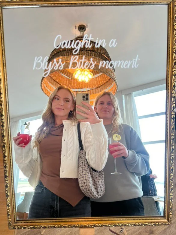 Two young women taking a selfie in front of a mirror during a social gathering, holding drinks, with a decorative ceiling light and a window in the background.