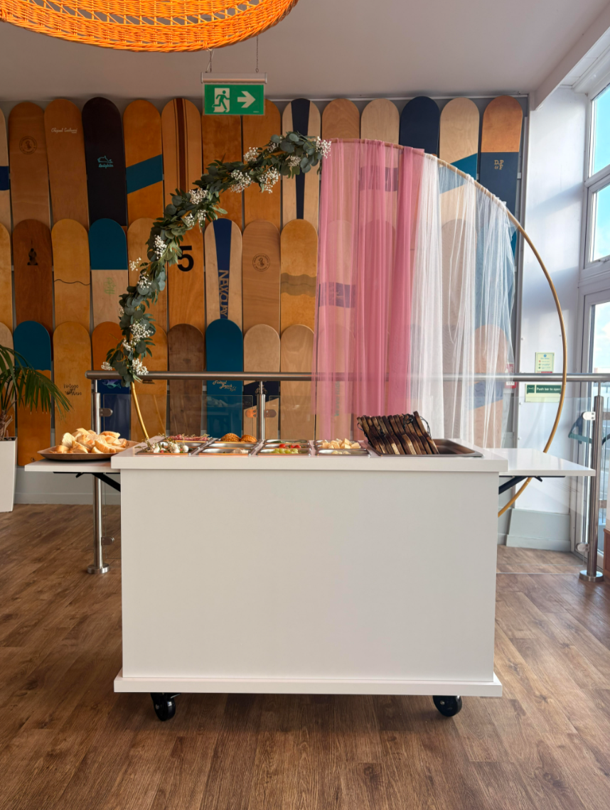A white mobile serving cart with food items in a cafe or restaurant. The background features a skateboard art wall, a hanging green exit sign, and a colorful circular art installation with pink, white, and blue sheer curtains.