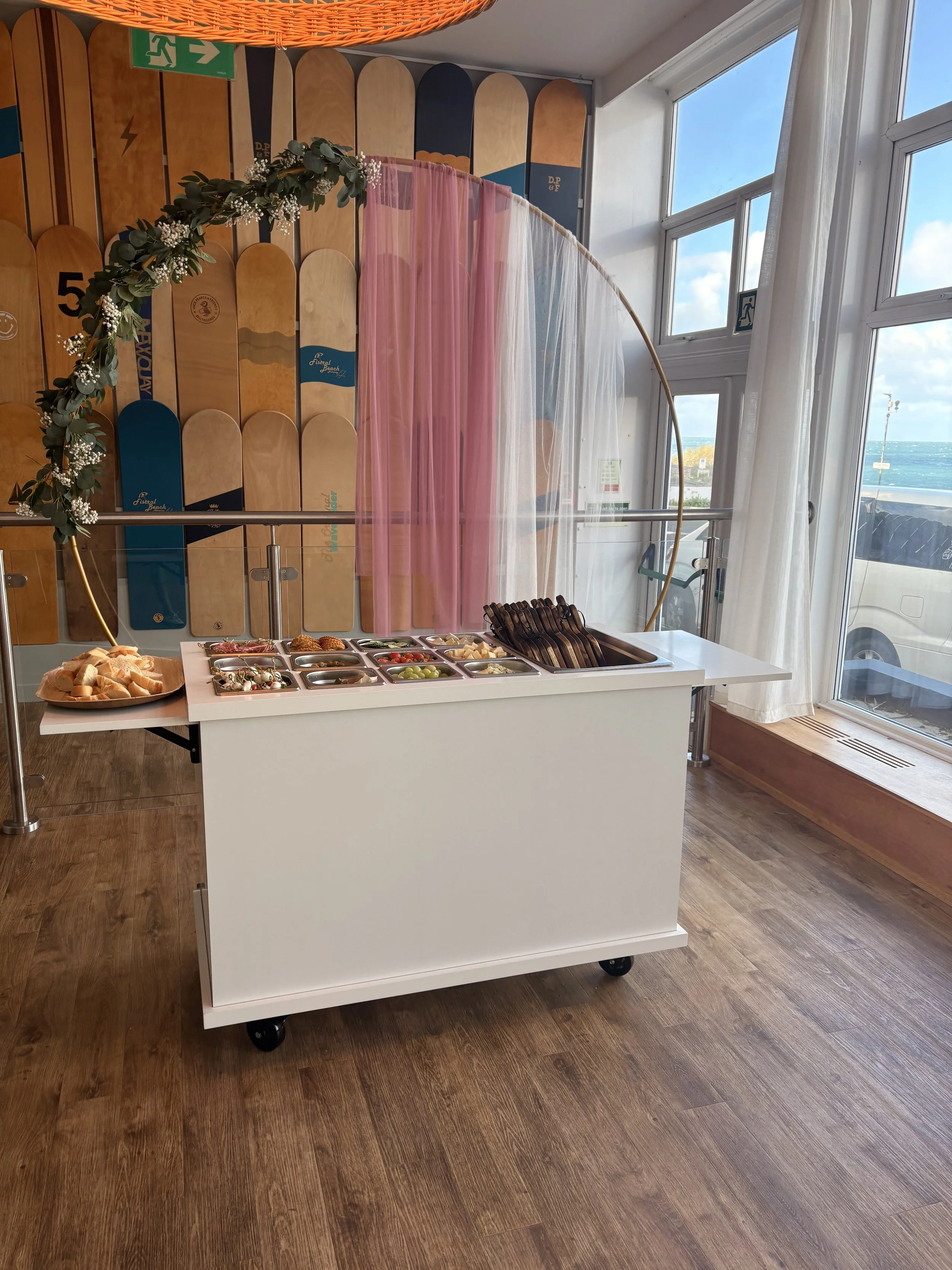 A buffet table with a variety of small dishes and bread, set against a wall with skateboard decks and pink and white curtains near large windows overlooking the ocean.