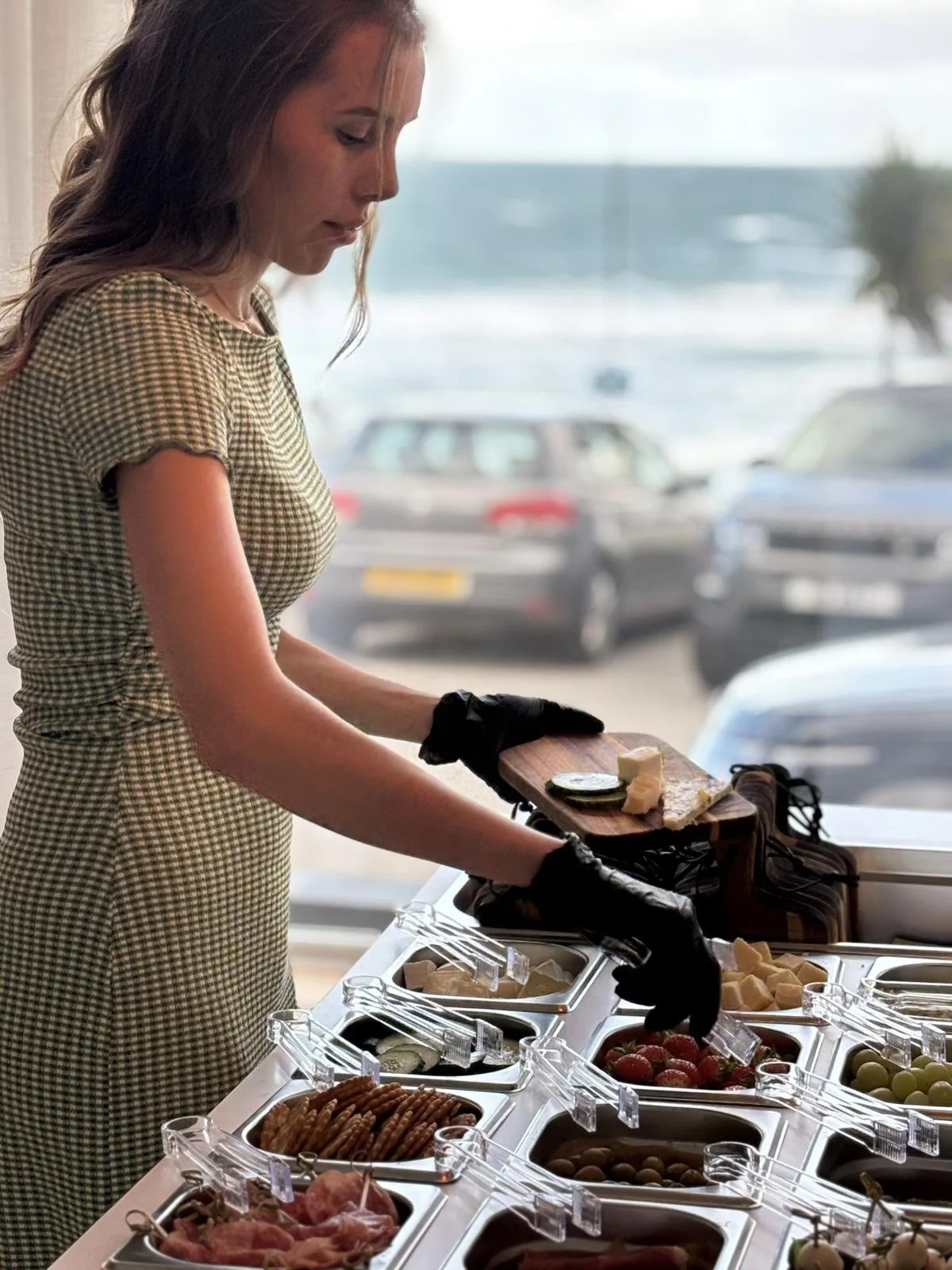 A woman in a checkered dress and black gloves serving herself food from a buffet with various food containers, including cheese, strawberries, grapes, meats, and vegetables, near a window overlooking parked cars outside.
