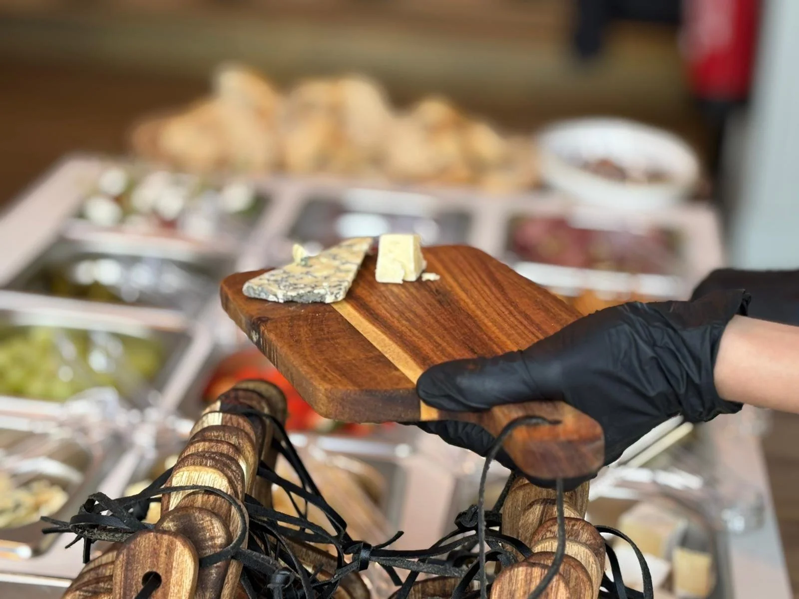 A person wearing a black glove holding a wooden cheese board with two pieces of cheese on it, in front of a display of various cheeses and toppings at a cheese shop.