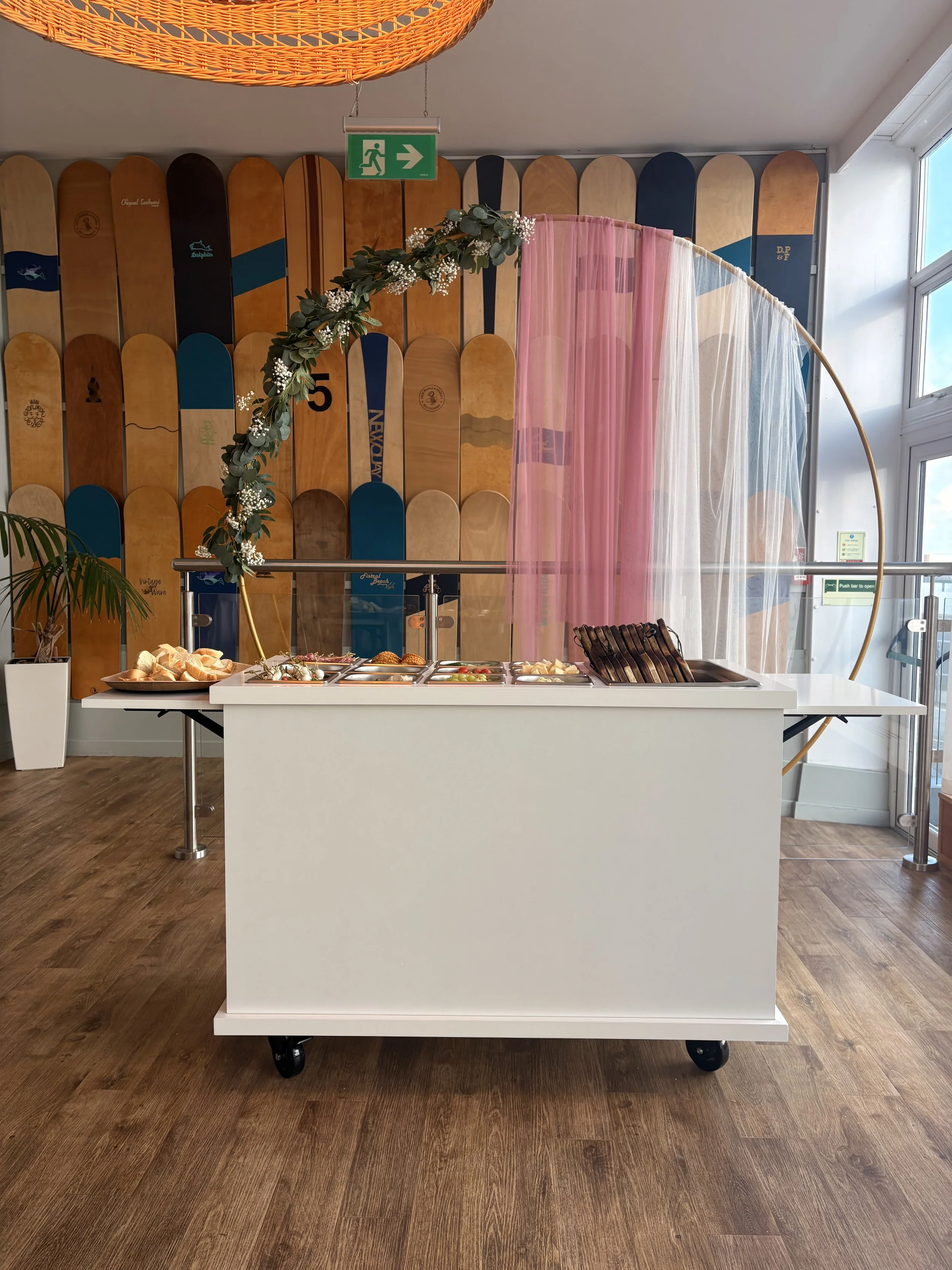 A white mobile serving cart with a variety of food and drink items, set against a background of skateboard decks and pink and sheer curtains, with a green exit sign above.