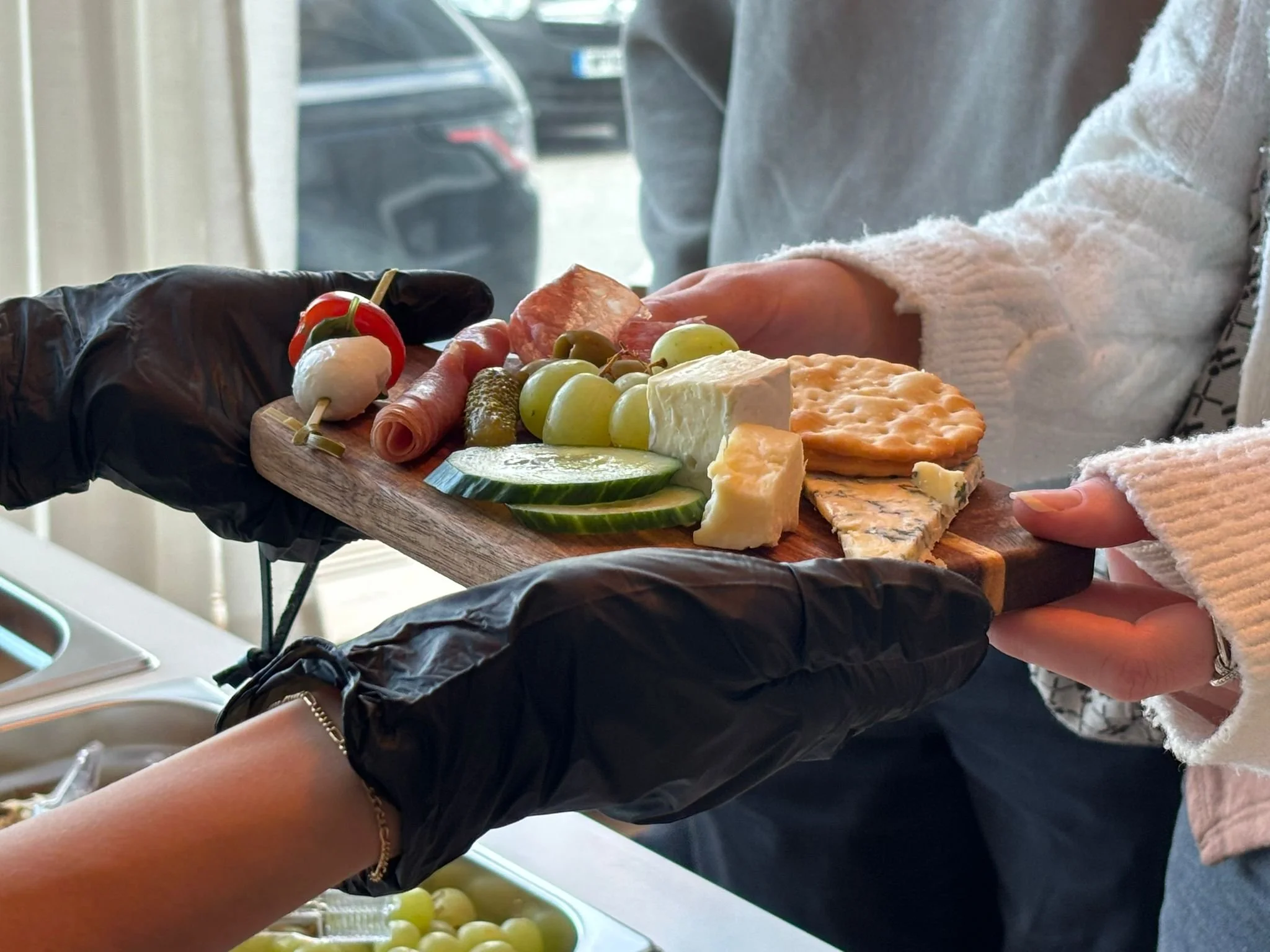 People exchanging a cheese platter with grapes, meats, cheeses, cherry tomato, cucumber slices, and crackers in a well-lit indoor setting.