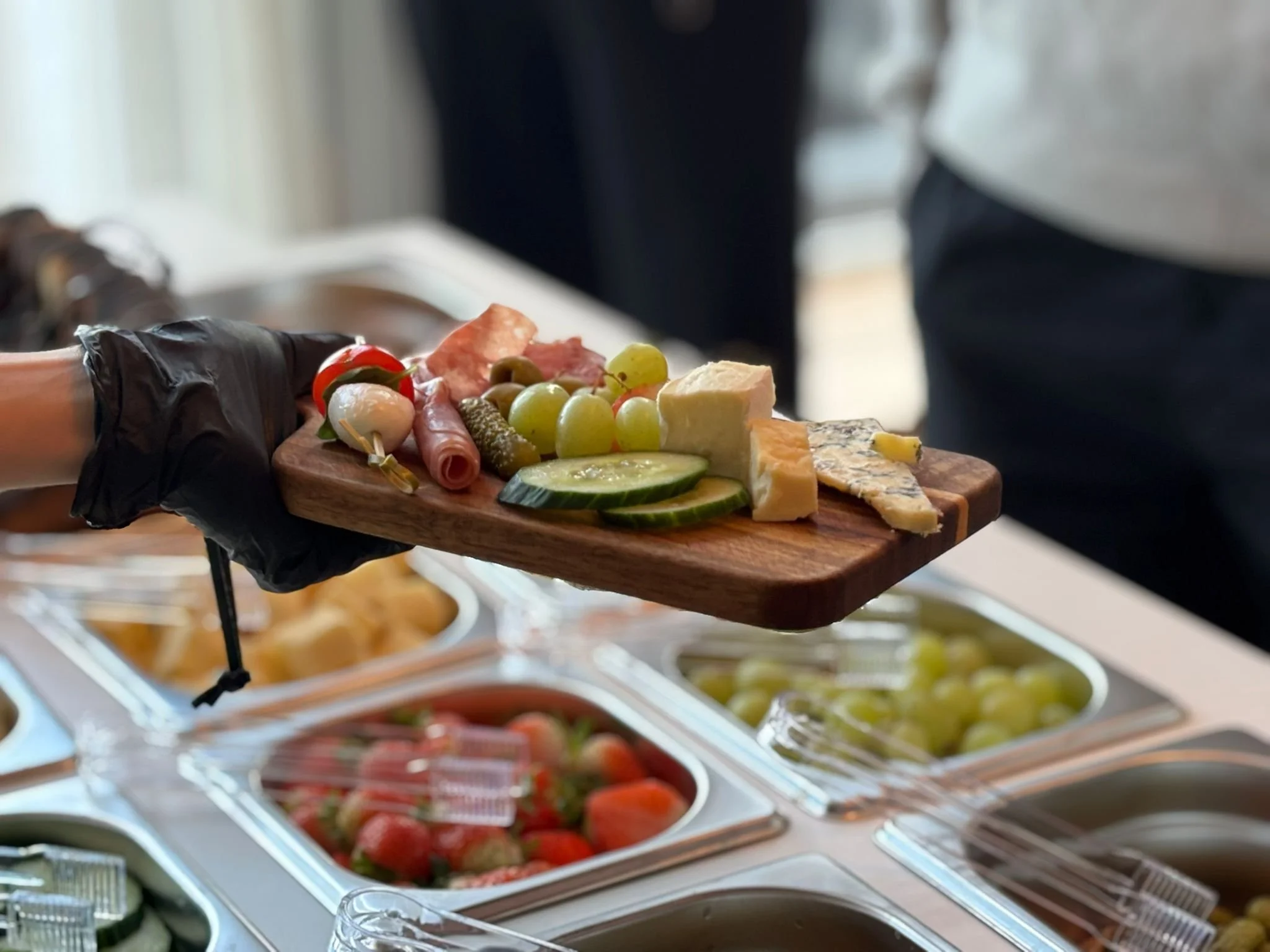 Close-up of a hand wearing a black glove holding a wooden platter with assorted cheese, green grapes, cherry tomatoes, cucumber slices, and cured meats, at a buffet or party setting.