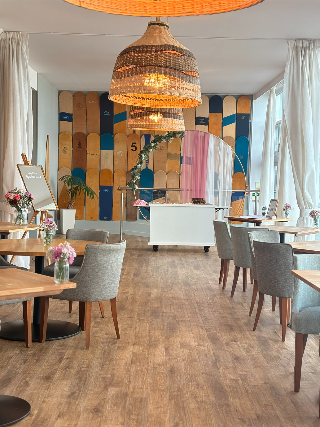 A cozy, well-lit cafe with wooden tables and chairs, decorated with small pink flowers in glass vases, a skateboards art wall in the background, and a white bar table in the center with a pink and white sheer curtain.