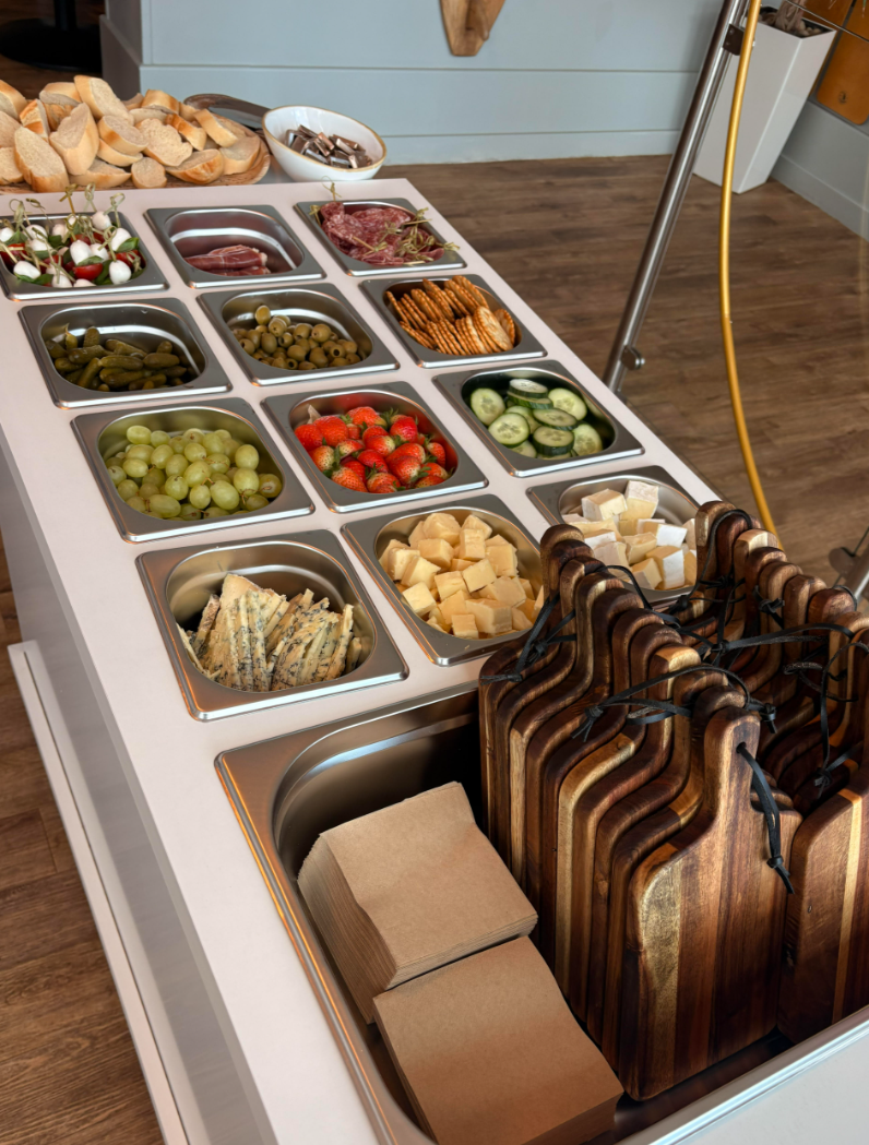 A charcuterie and cheese platter with various fruits, vegetables, meats, cheeses, crackers, and pickles on a white table, with stacked cutting boards and napkins.