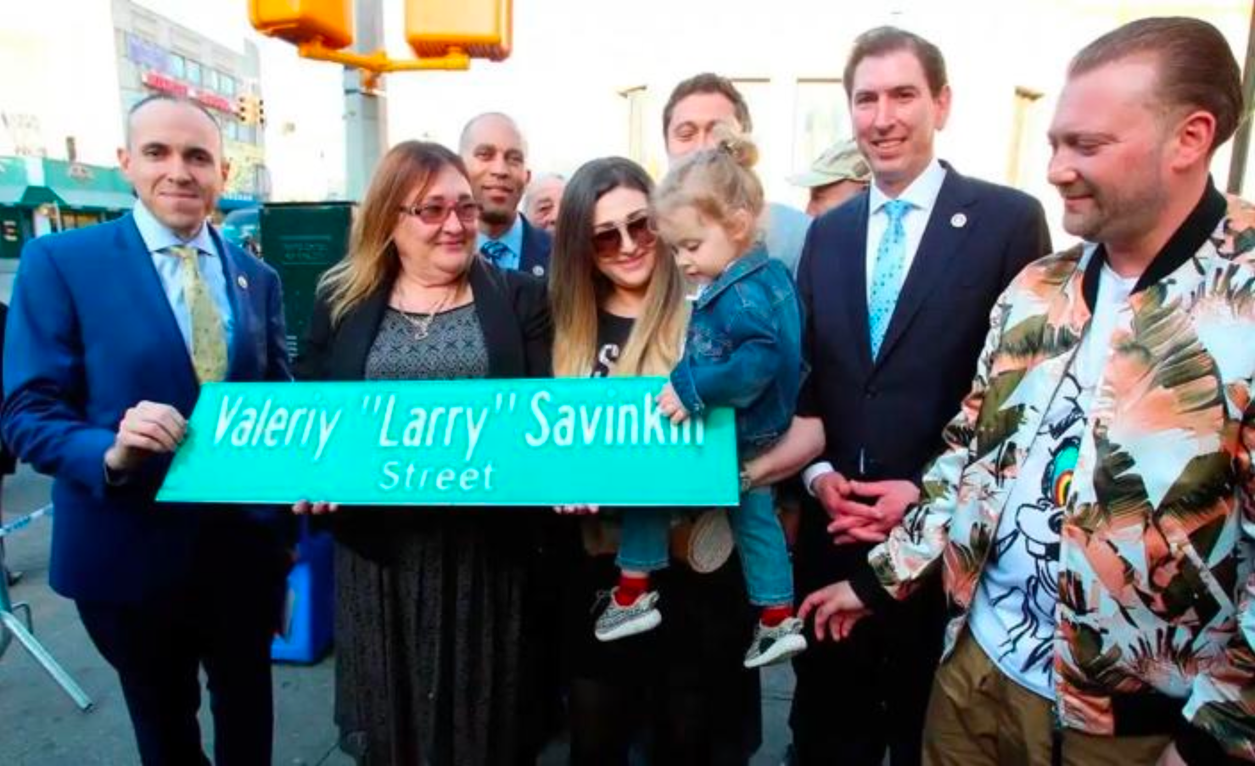 Group of people gathered around a street sign that reads 'Valery Larry Savinkoff Street'. The group includes men and women, some with sunglasses, and a young girl being held by a woman. They are outdoors in daylight, possibly at a street naming ceremony or community event.