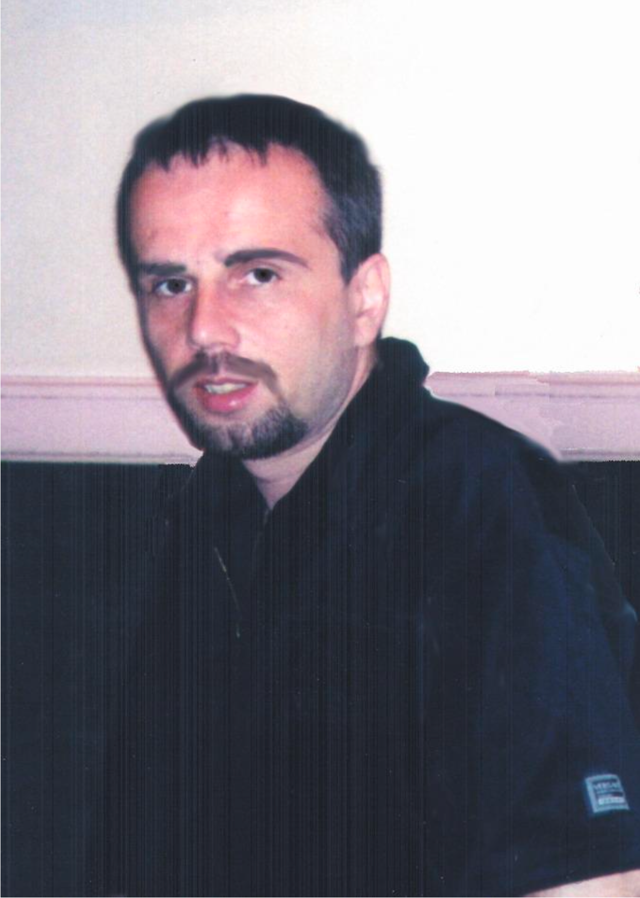 A man with short dark hair, a goatee, and wearing a black hoodie, sitting against a blank white wall.