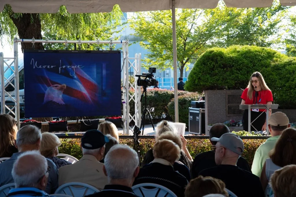 A woman giving a presentation on an outdoor stage with a screen displaying the American flag and the words "Never forget." Audience members are seated in front, watching outside in daytime.