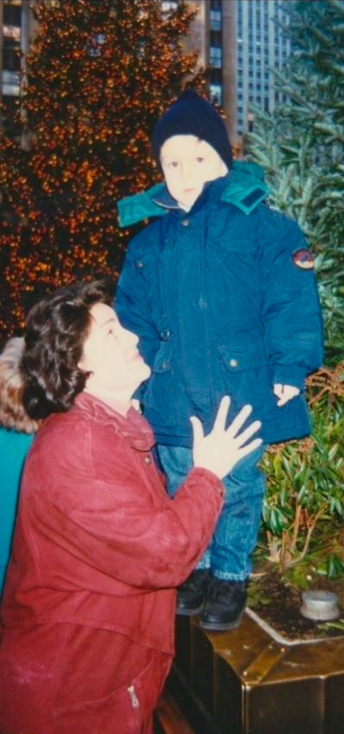 A woman with short curly hair and a red jacket kneeling with her hand on a child's chest, looking at him. A boy in a blue winter coat and black beanie standing in front of a decorated Christmas tree with orange lights, in an outdoor urban setting with tall buildings and greenery.