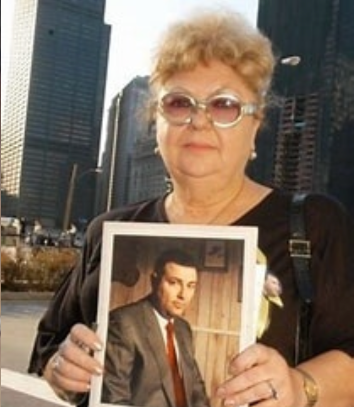 An elderly woman with short curly hair and glasses holding a framed photograph of a man in a suit, outdoors in an urban setting with tall buildings.