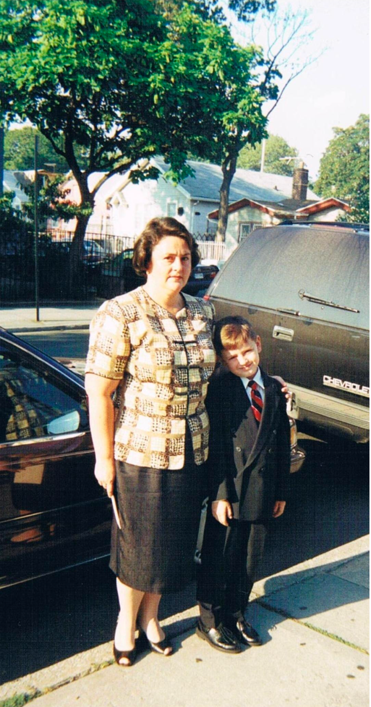 A woman and a young boy standing on a sidewalk next to a black car and a gray Chevrolet vehicle, with trees and houses in the background on a sunny day.