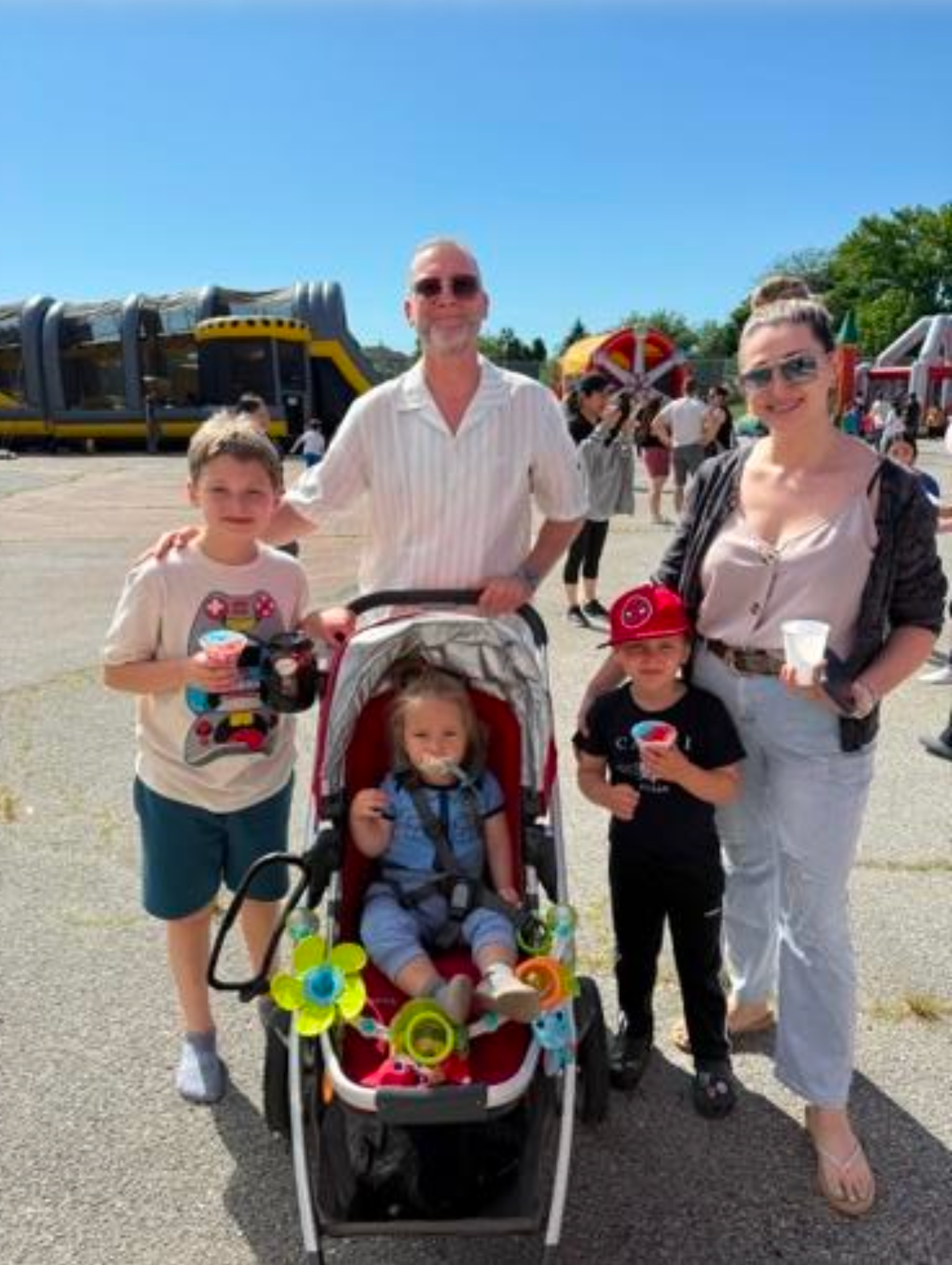 A group of six people, including children, standing outdoors at an amusement park or fair on a sunny day. There are rides and other visitors in the background.