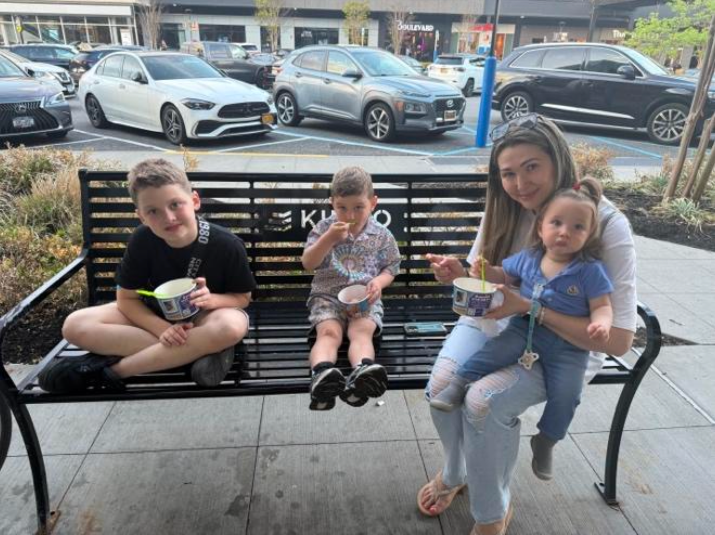 Four children sitting on a black metal outdoor park bench eating ice cream, with a woman sitting on her knees beside them. They are outside near a parking lot with several cars and a shopping mall in the background.